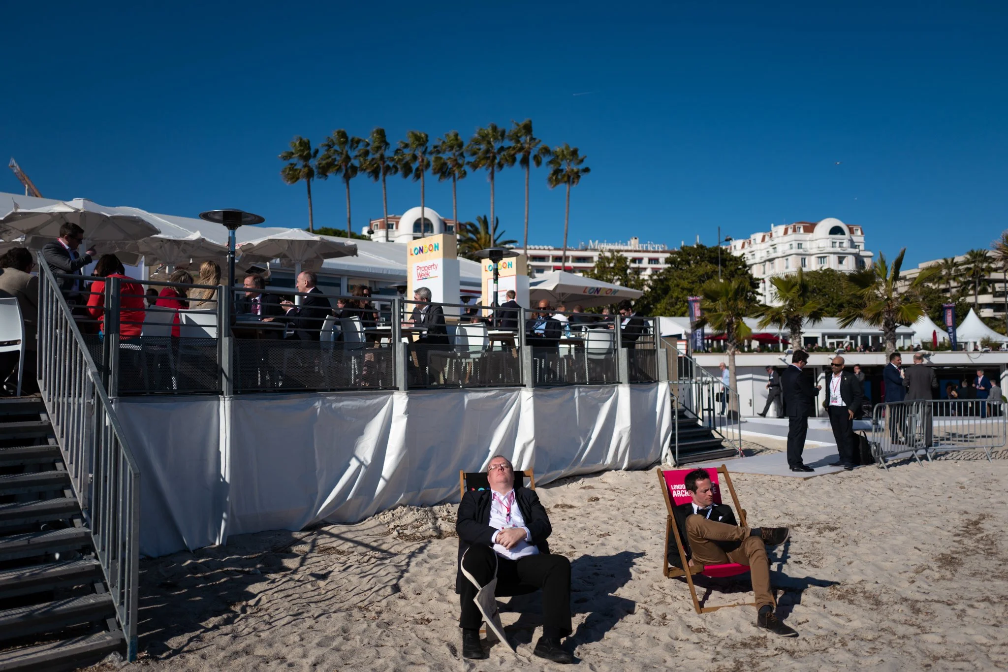 Beach scene with two men relaxing on deck chairs on the sand, people socializing under umbrellas on a raised platform, tall palm trees, and buildings in the background on a sunny day.