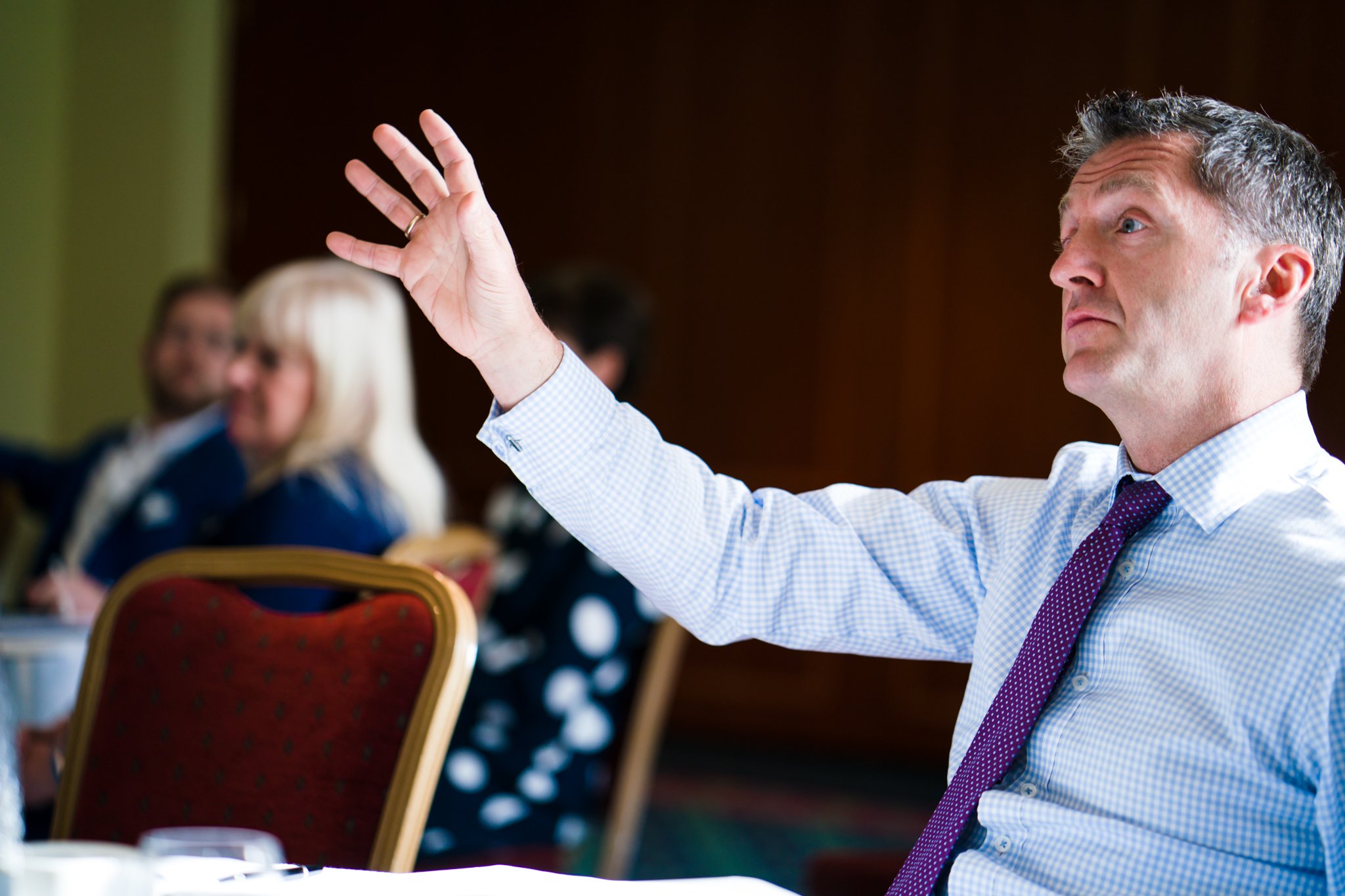 A man in a dress shirt and tie raising his hand during a meeting or conference, seated at a table with other people in the background.