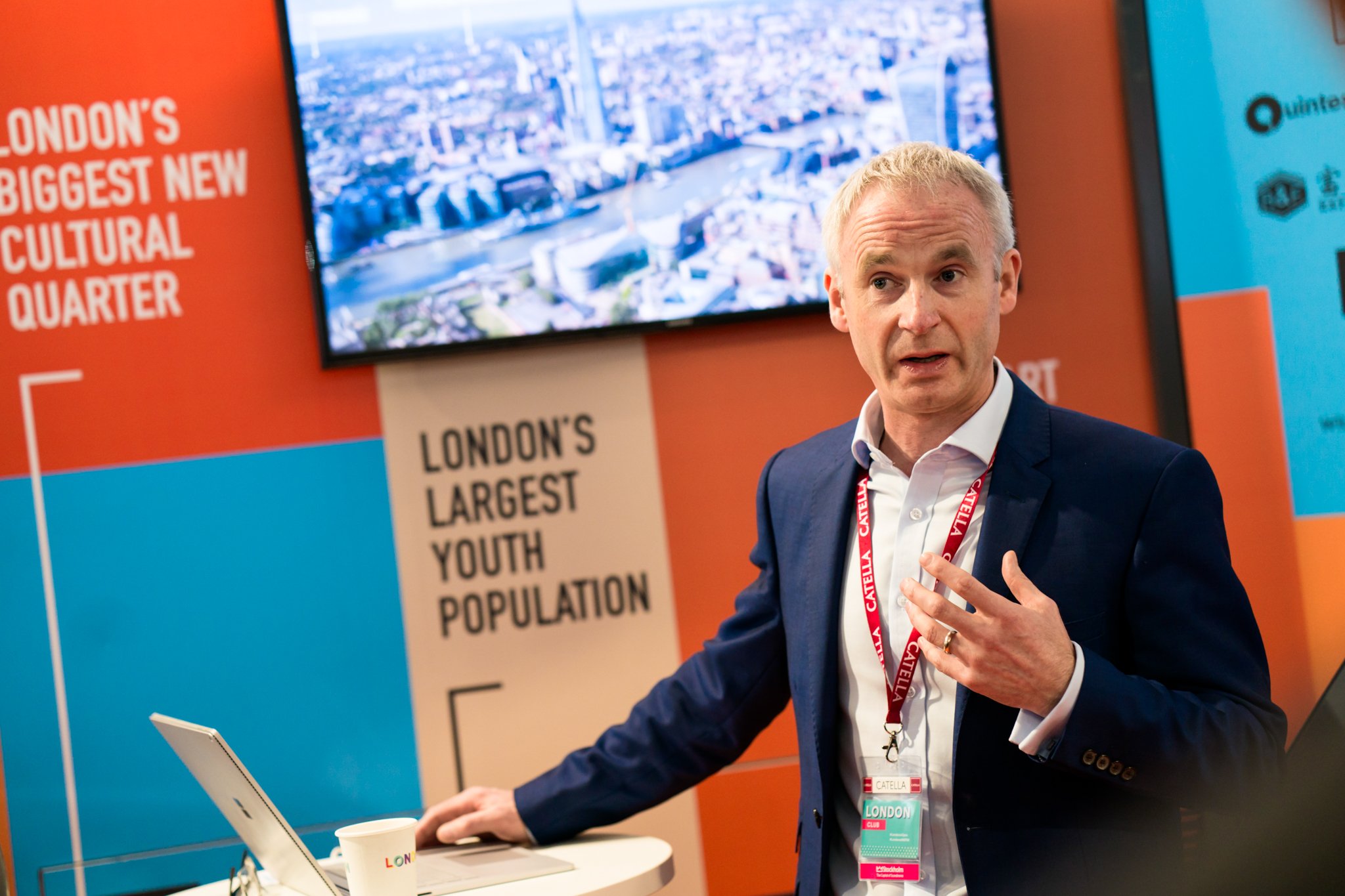 A man in a navy suit and white shirt speaks at a conference booth with banners that read 'London's Biggest New Cultural Quarter' and 'London's Largest Youth Population.' He has a red conference badge around his neck and is gesturing with his hand whi