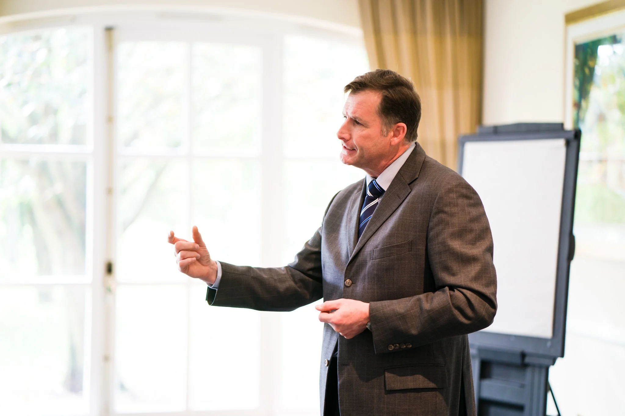 Man in a gray suit giving a presentation in a bright room with large windows and a whiteboard.
