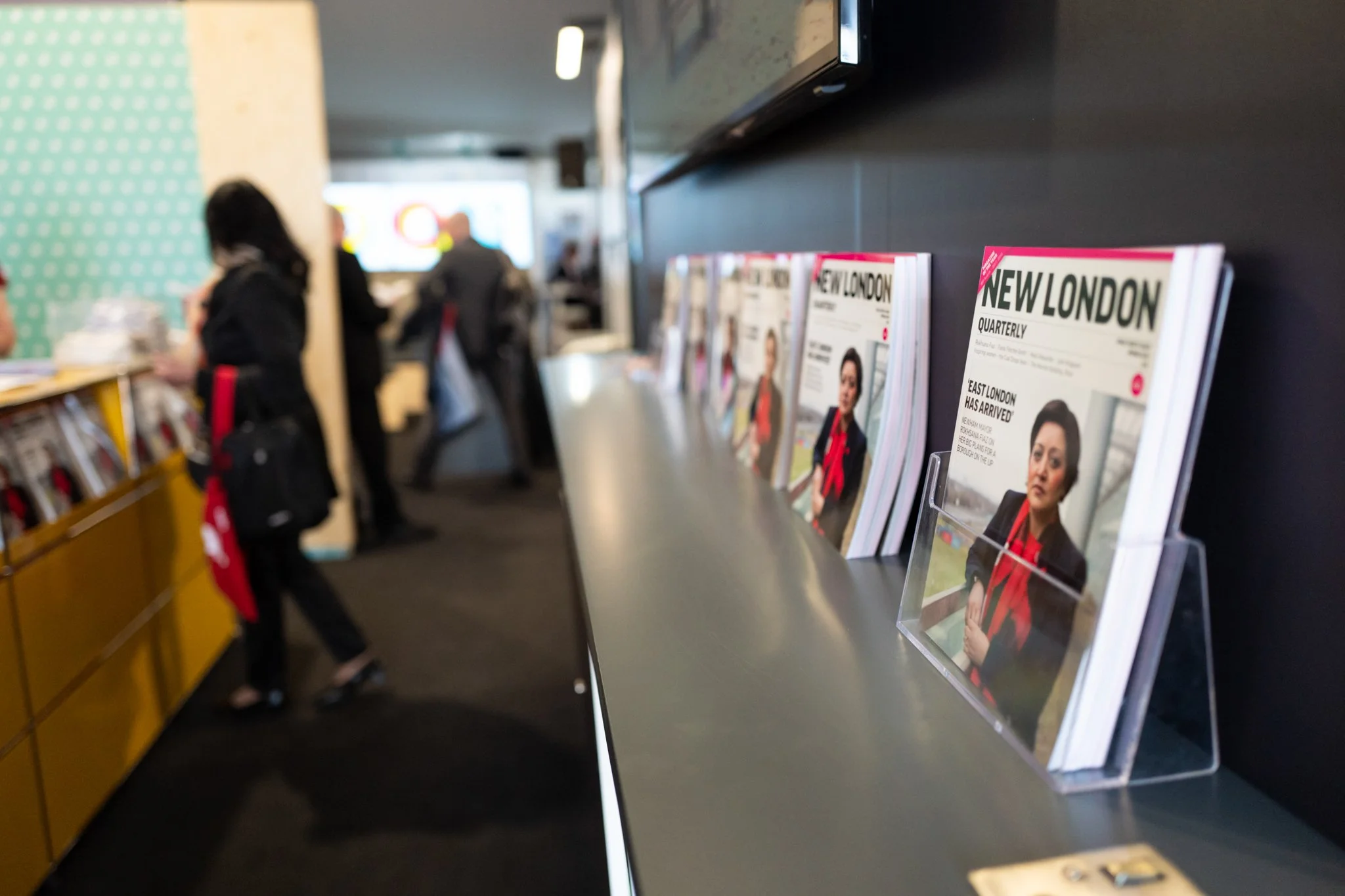 Brochures or magazines titled 'New London' displayed on a shelf in a public space, with people standing and walking in the background.