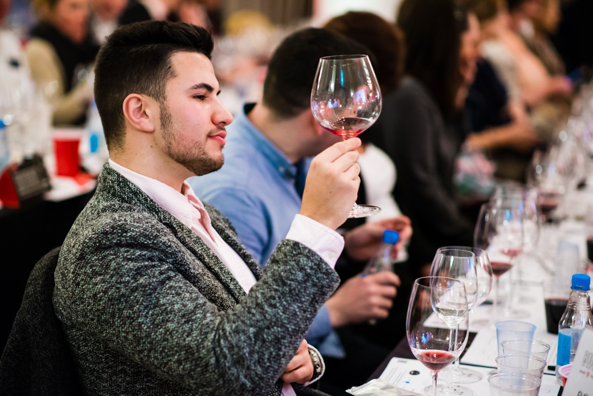 A young man in a gray sweater tasting wine at a wine tasting event, holding a wine glass up to examine it among other people at the table.