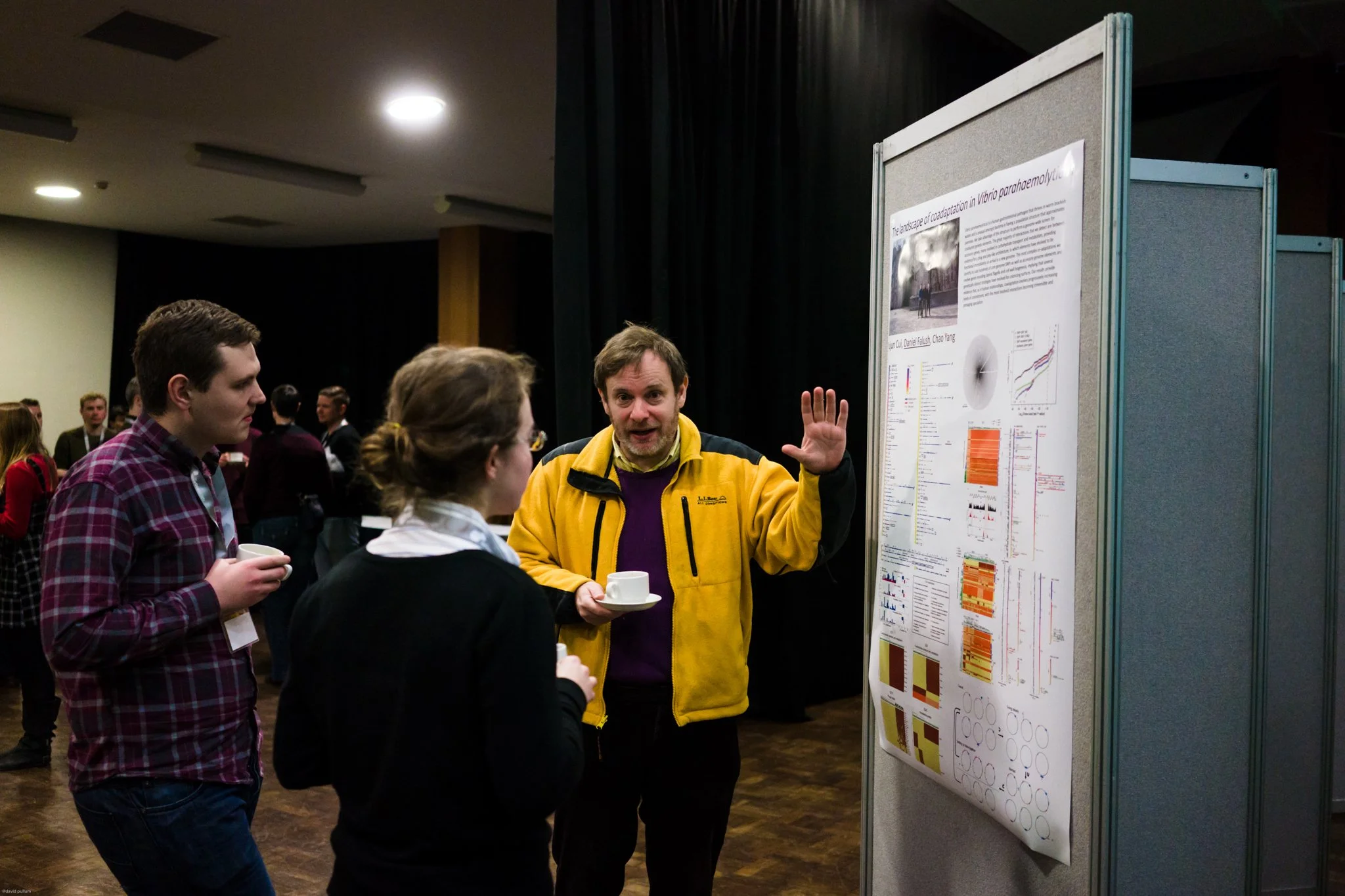 A man in a yellow jacket describing a scientific poster to two people at a conference.