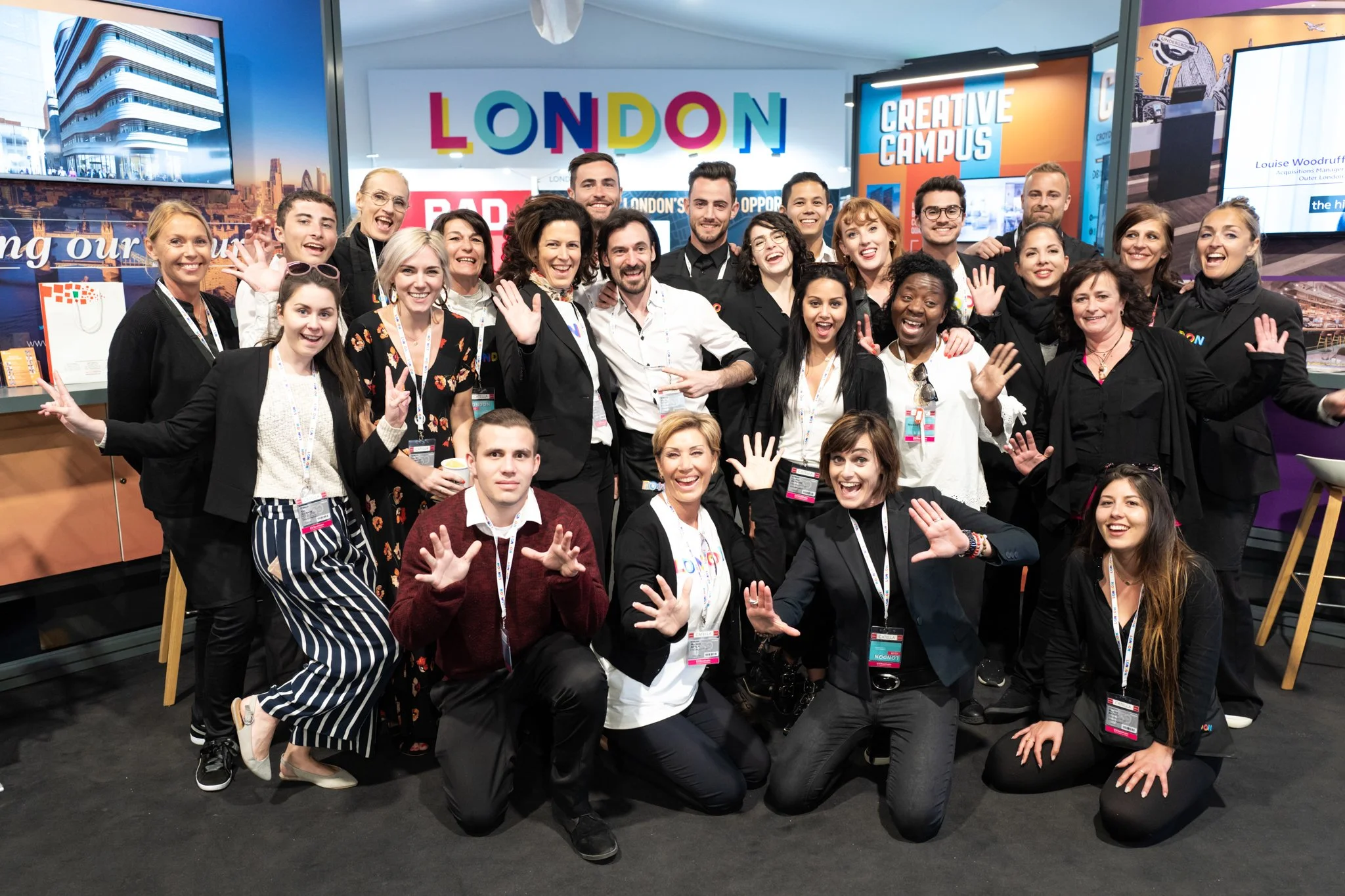 A group of smiling people at a conference or event, wearing badges and making cheerful gestures. Behind them, colorful signs with the words 'LONDON', 'CREATIVE CAMPUS', and other text, along with screens displaying various images and graphics.