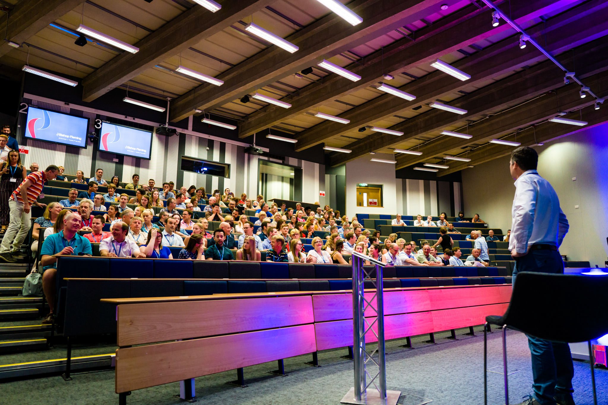 A man in a white shirt stands at a podium in front of an audience seated in a lecture hall with tiered seating and multiple large screens displaying a logo.