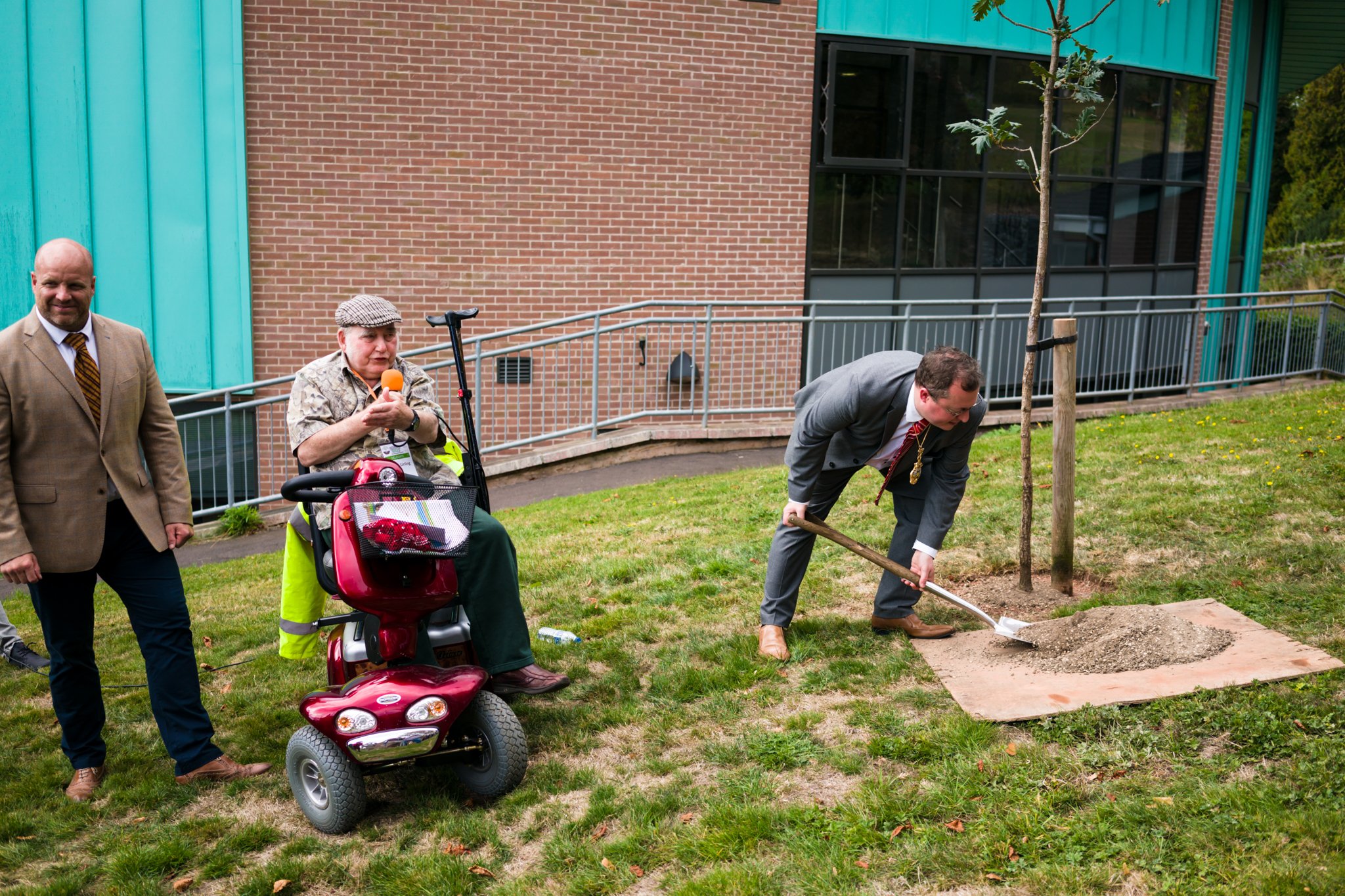 Men, including a man in a gray suit and tie, participate in a tree-planting event on a grassy area in front of a brick and teal-colored building. One man is shoveling dirt onto a newly planted tree. Another man, seated on a mobility scooter and dress
