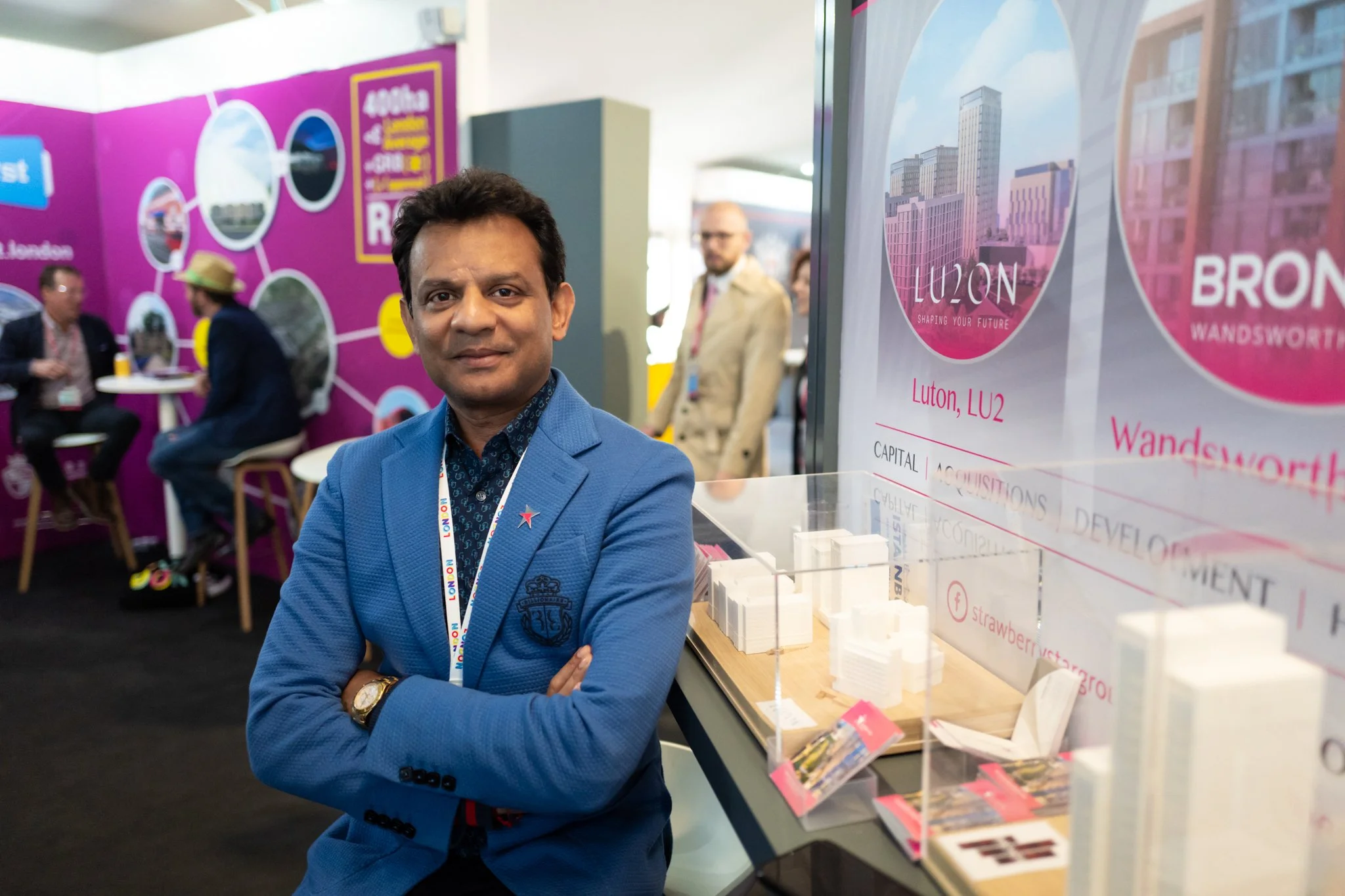 A man standing with arms crossed at a conference, wearing a blue blazer and a gold watch, with people and promotional displays in the background.