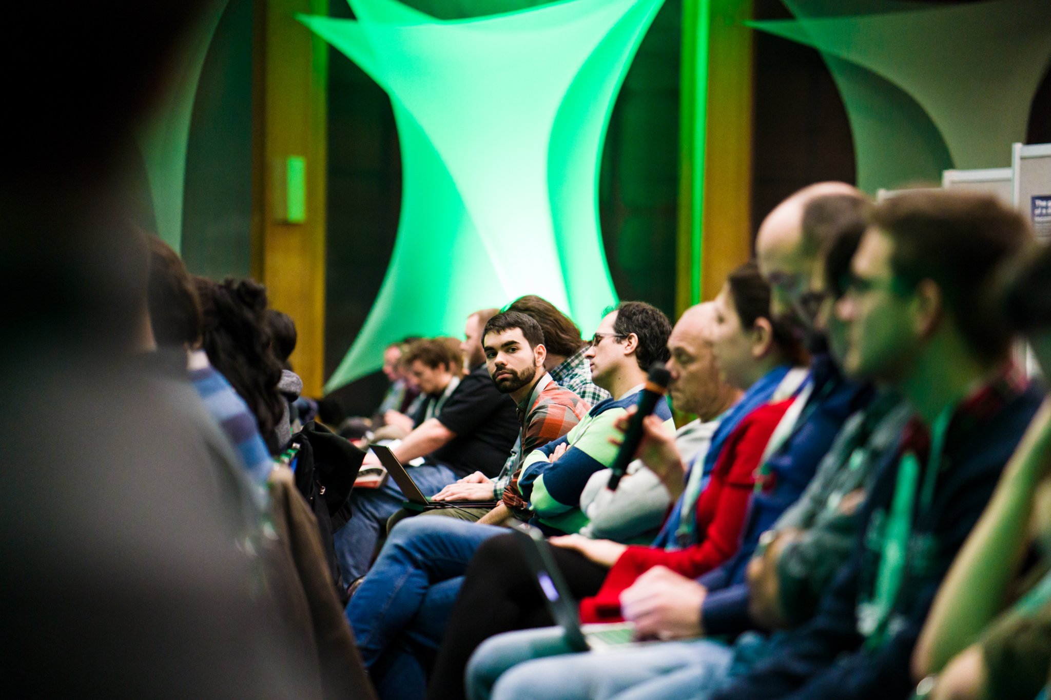 Audience of people sitting in rows at a conference or seminar, with one individual holding a microphone, in a room with green-lit abstract decorations on the walls.