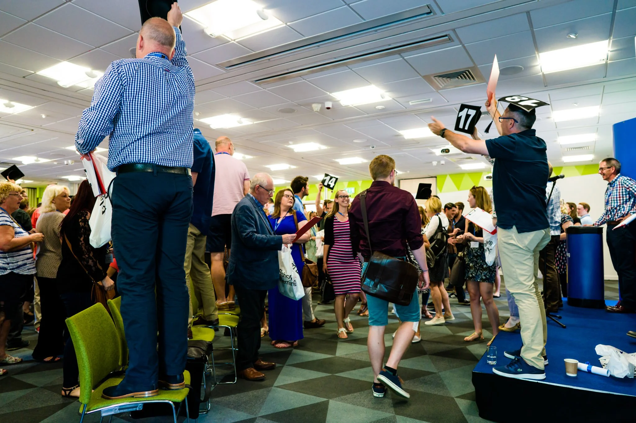 Crowd of people at an auction, with one man on stage holding up a bid card, others looking at paperwork or observing.