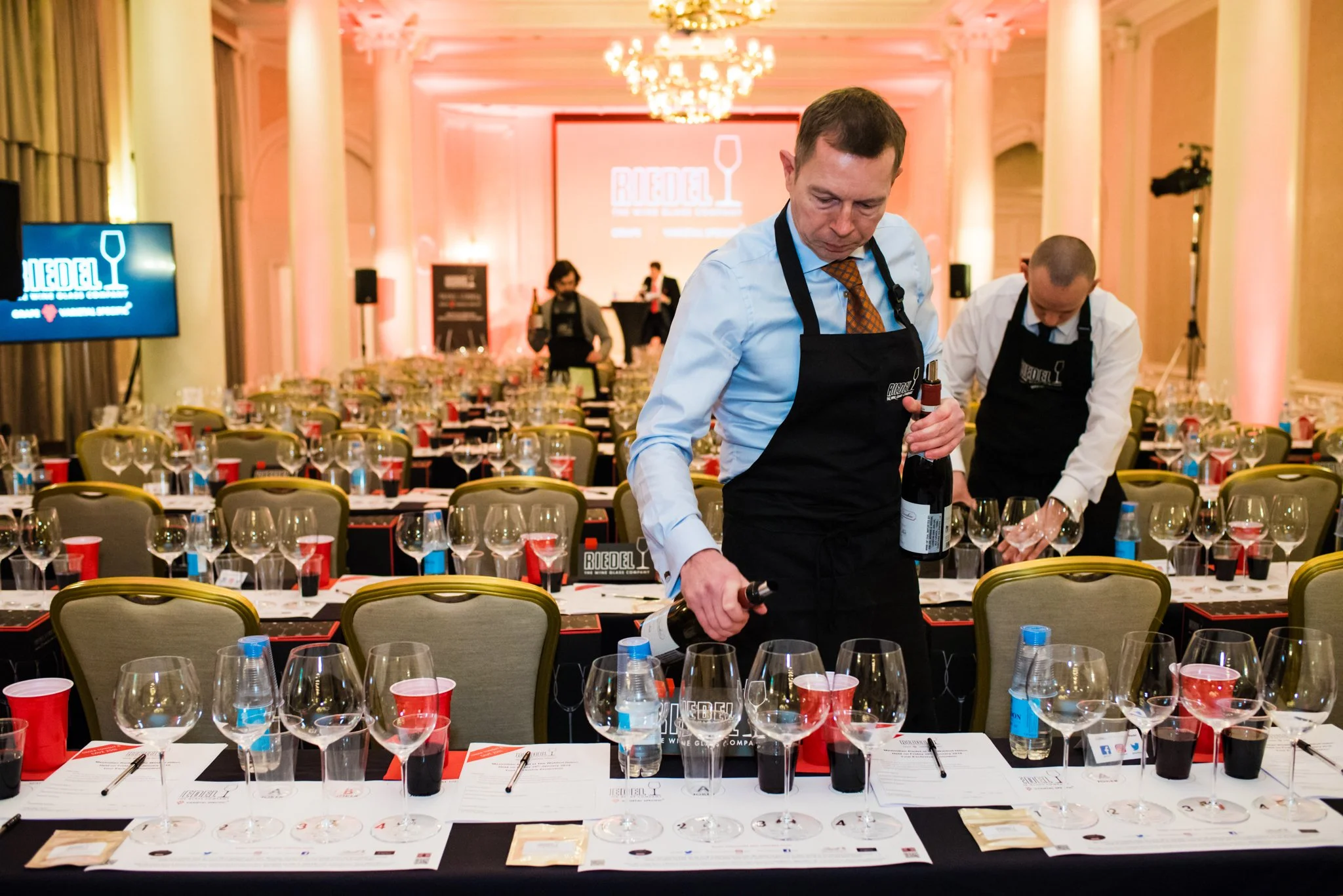 A man in a light blue shirt and tie, wearing a black apron, holds a wine bottle and a wine opener, preparing for a wine tasting event in an elegant, well-lit banquet hall with rows of tables set with multiple wine glasses, bottles of water, and tasti