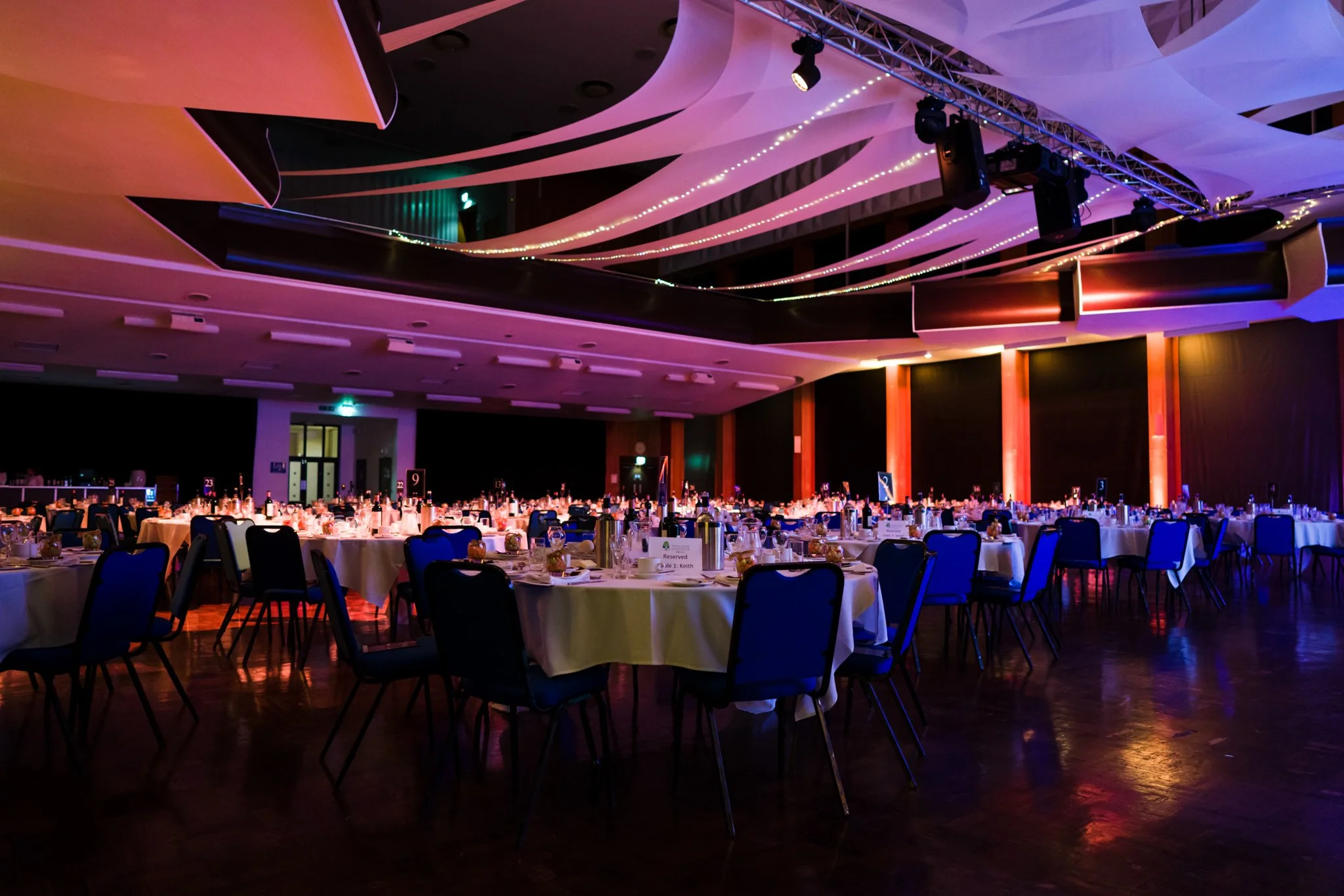 A large banquet hall decorated with colorful lighting, filled with round tables covered in white tablecloths, set for a formal event.
