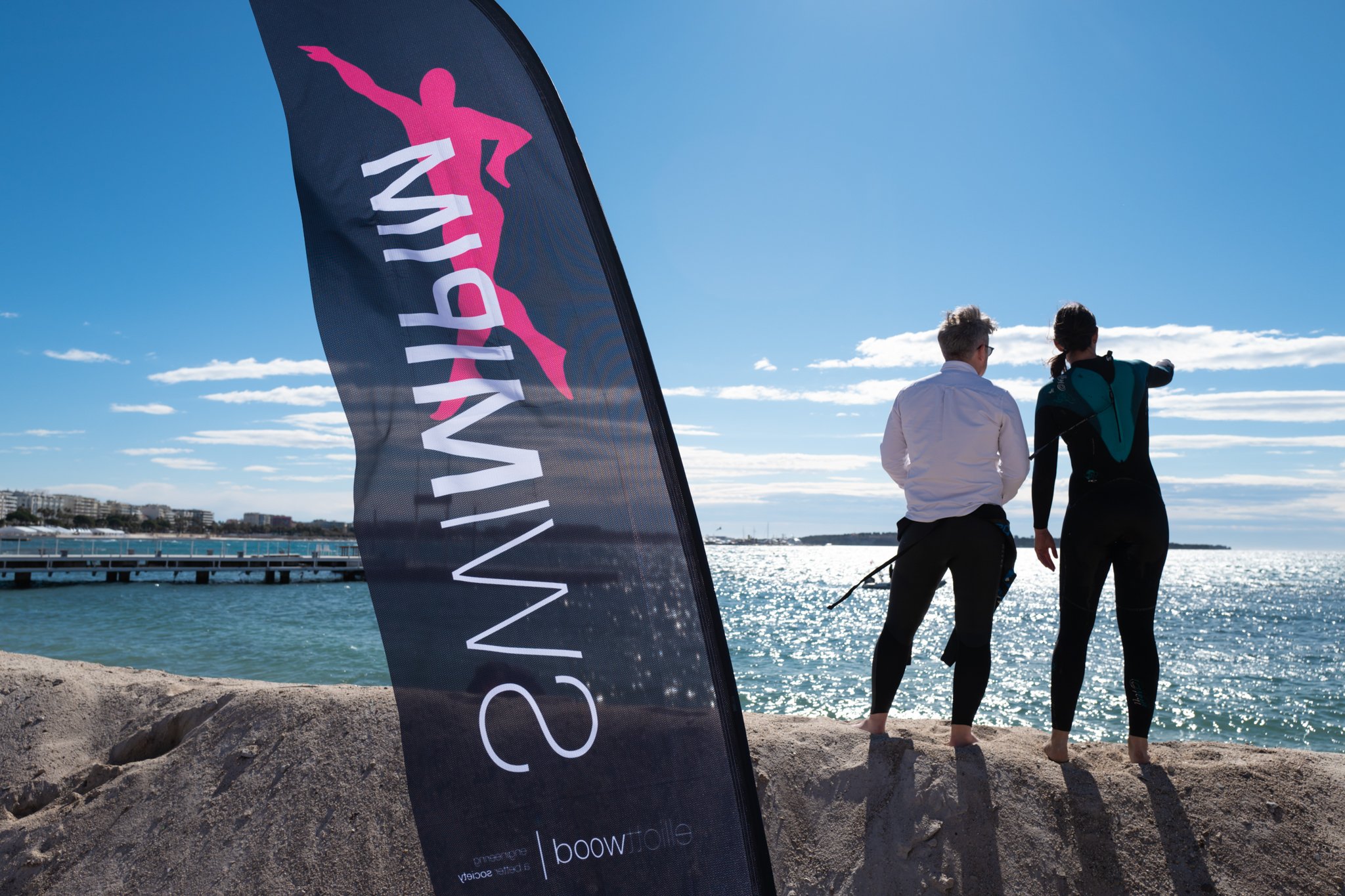 Two people in wetsuits standing on a sandy beach by the water, looking at the ocean on a sunny day with a blue sky and scattered clouds, with a flag reading 'SWIM SURF' in the foreground.