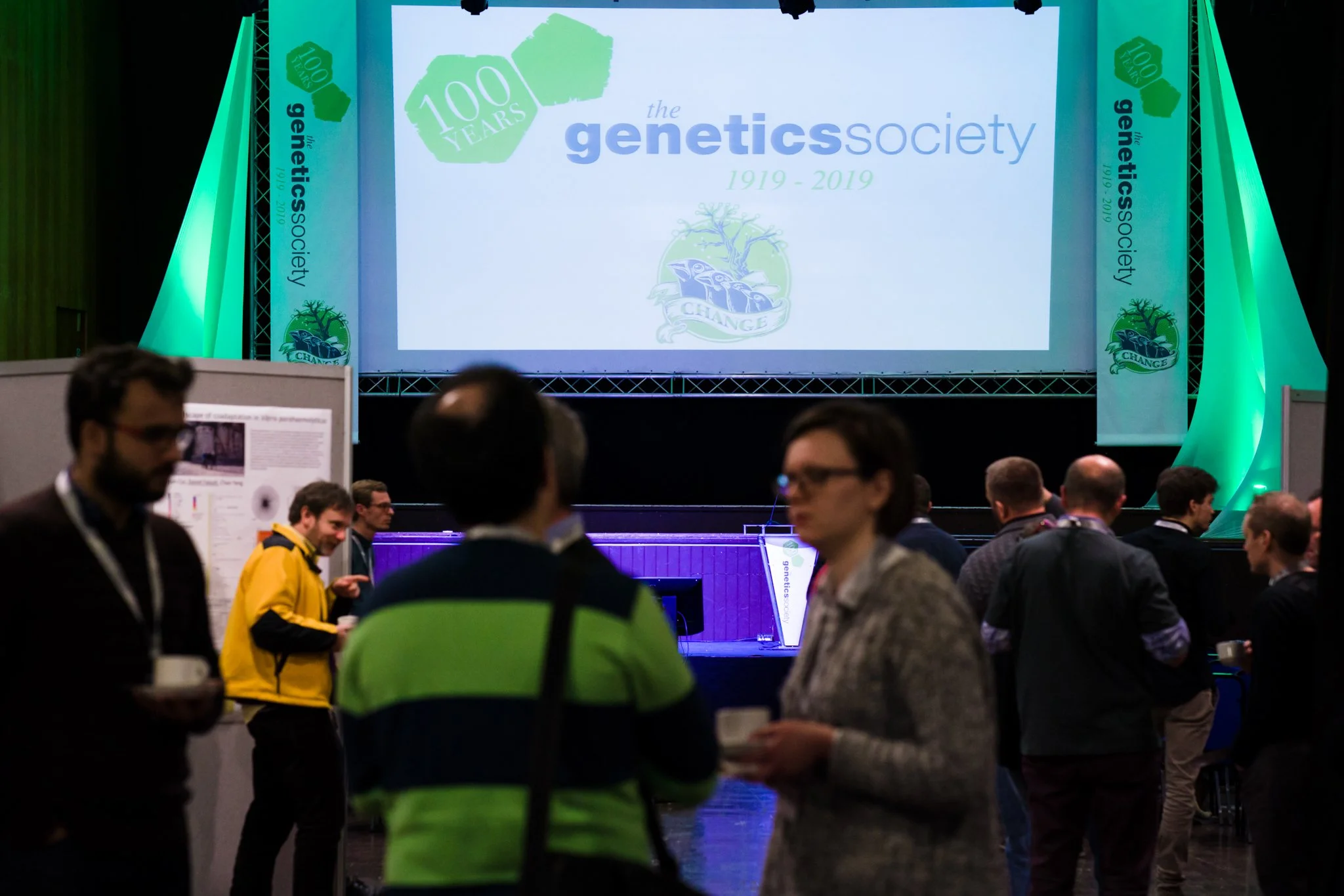 People attending a conference with a large screen displaying the 100th anniversary of the Genetics Society, 1919-2019, with a logo and decorative banners.