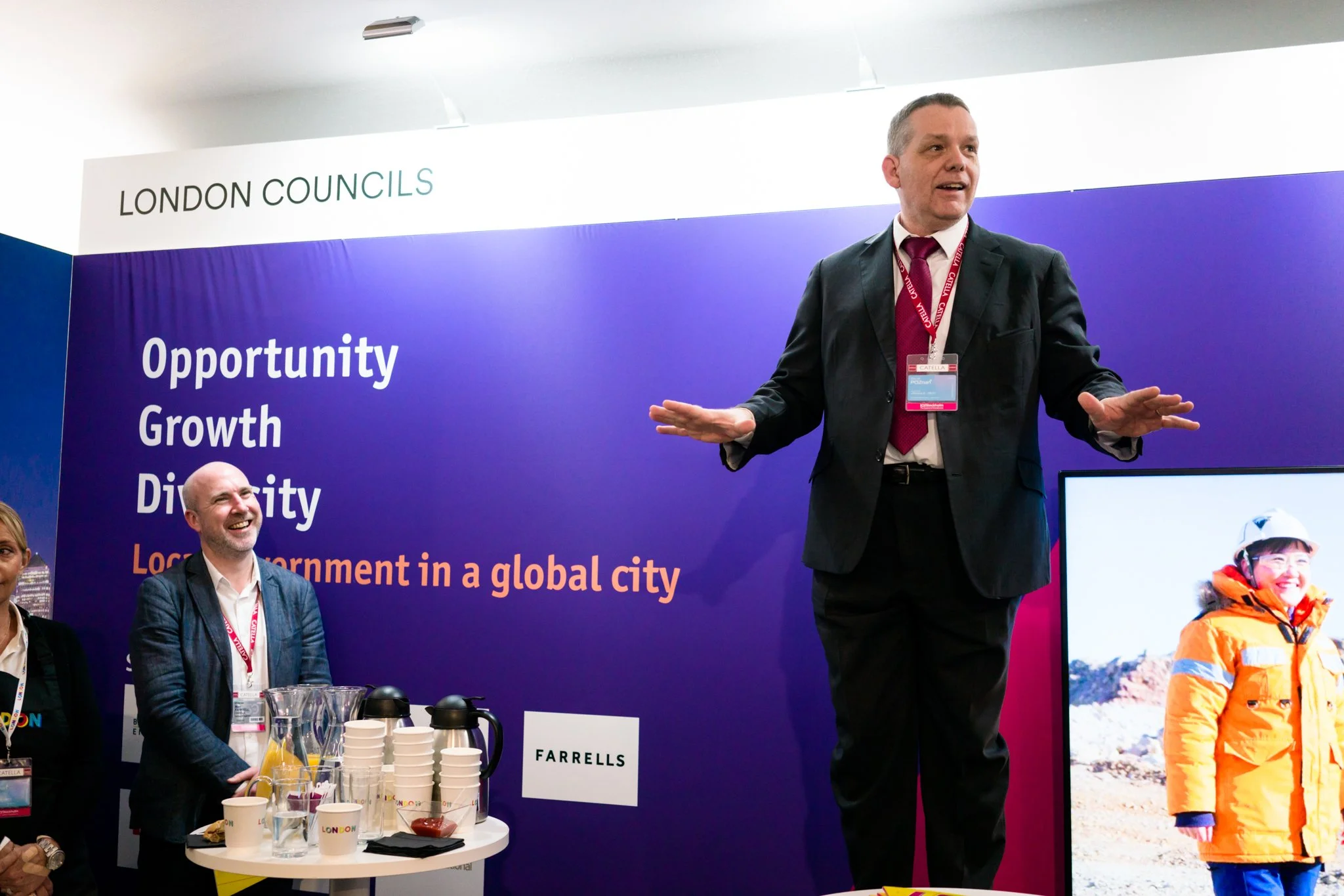 A man in a suit speaking with his hands outstretched at a conference booth with a blue and purple background. The background features the words "Opportunity Growth Dignity" and mentions "London Council" with a smaller sign saying "Farrells." There ar