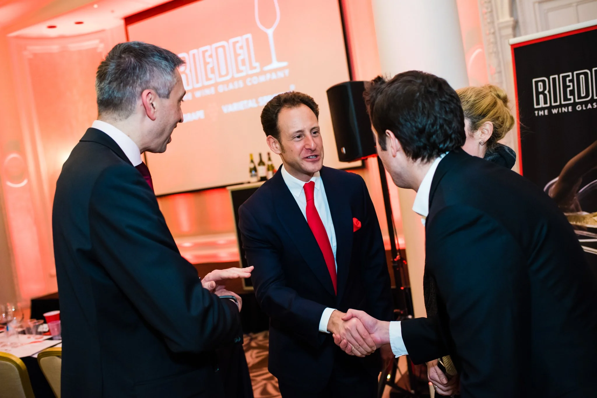 Four people in business attire are at a formal event, engaging in conversation. One man in the center, wearing a navy suit and a red tie, is shaking hands with another man dressed in a black suit. A large screen with the 'Riedel' logo and a black and