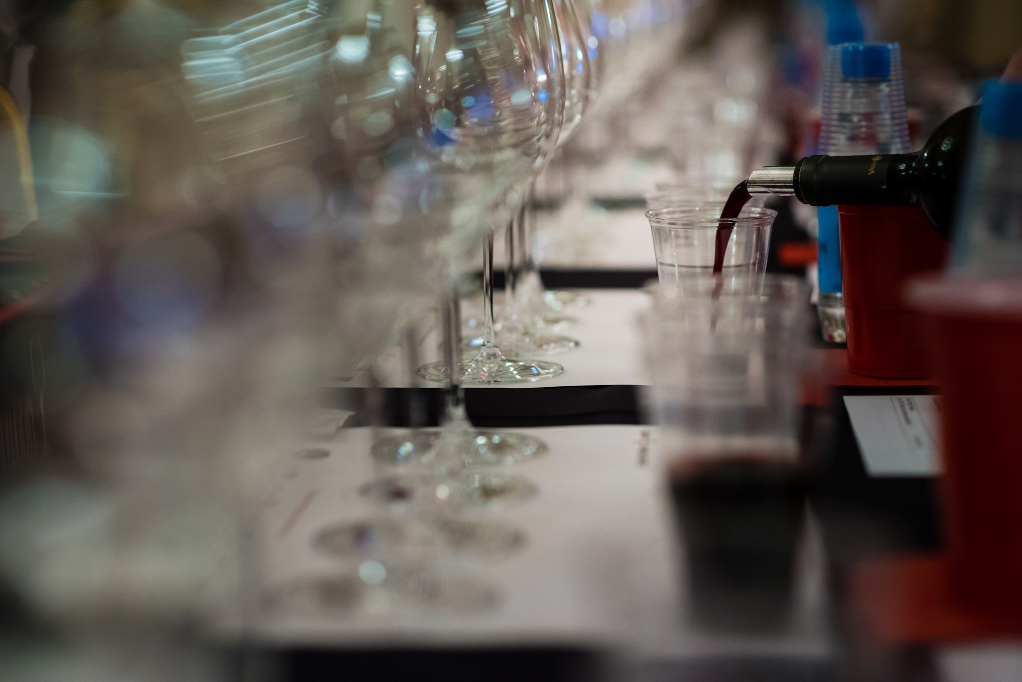 Wine being poured into a plastic cup at a bar or table with glassware and water bottles in the background.