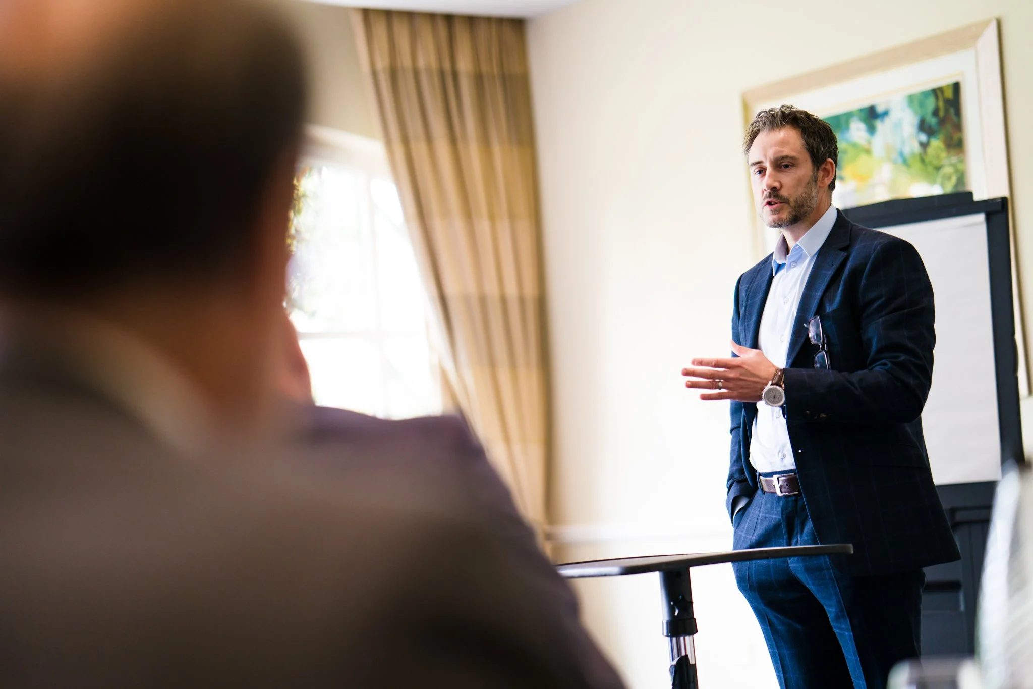 A man in a checkered suit giving a presentation in a room with a whiteboard, while an audience member in the foreground listens.