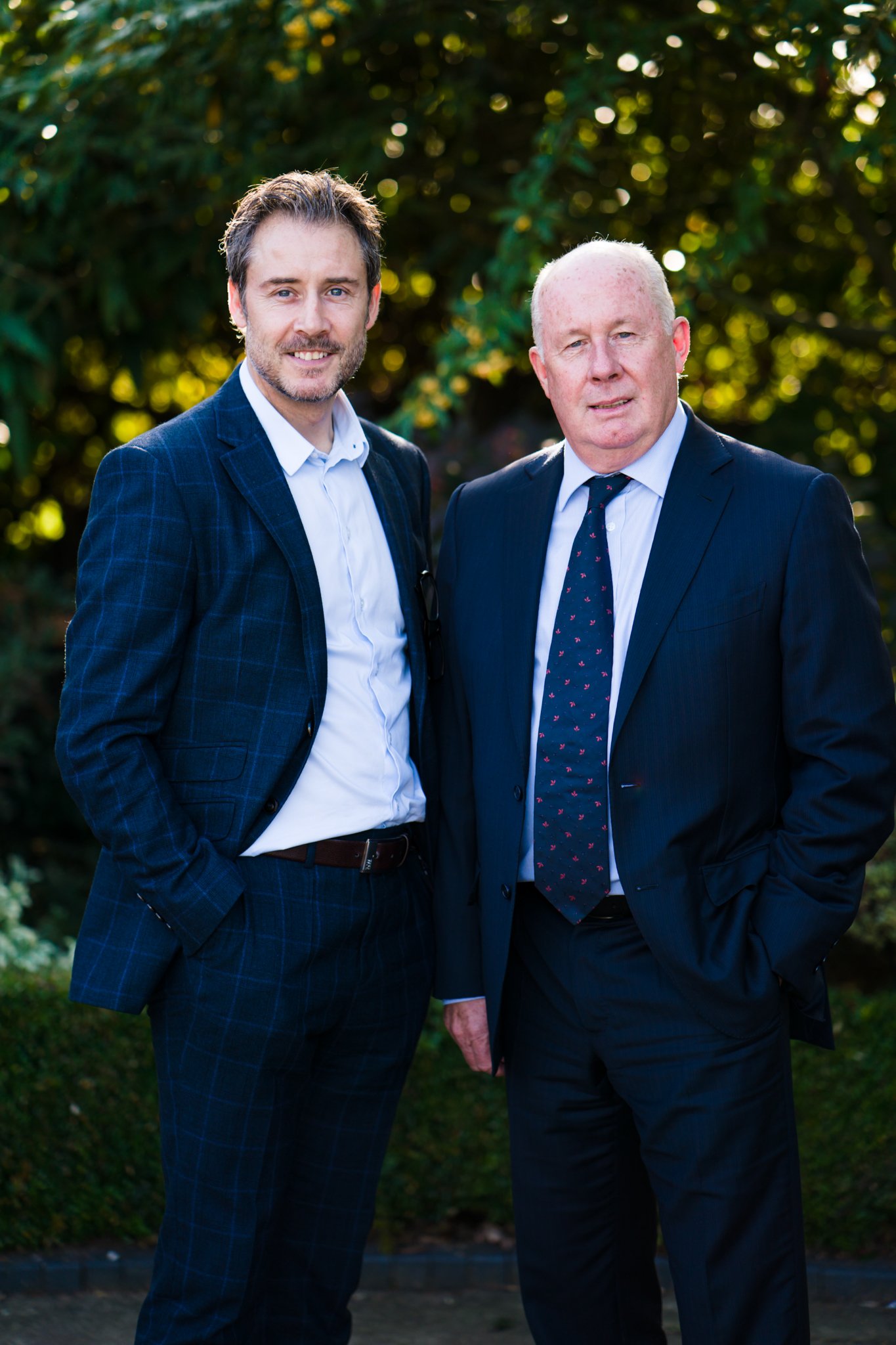 Two men in suits standing outdoors in front of greenery, smiling.