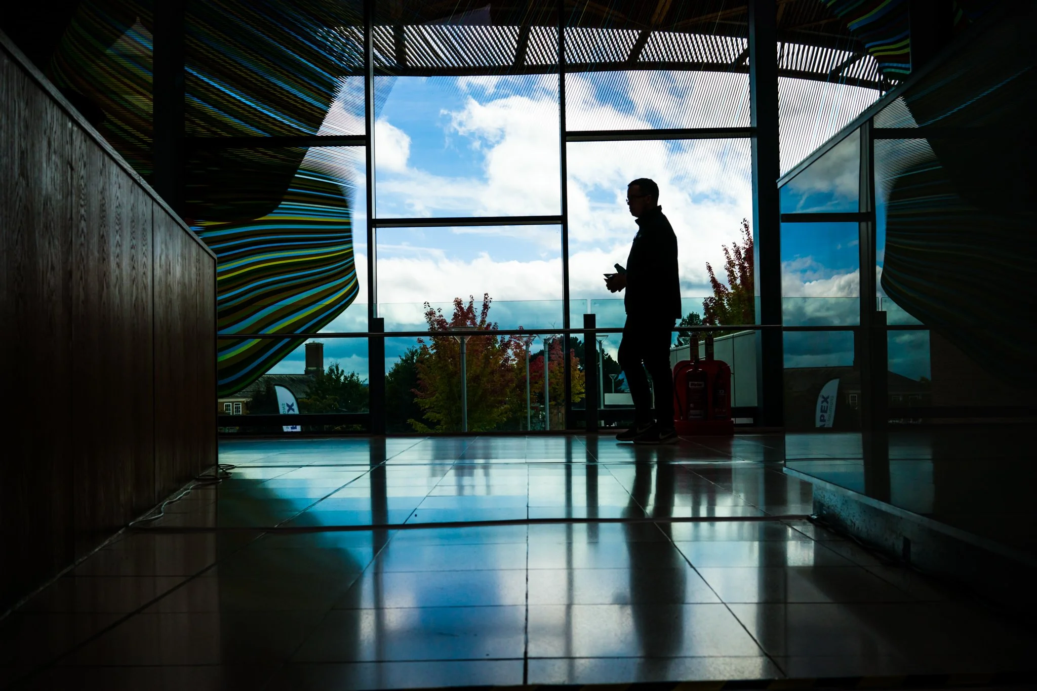 Silhouette of a man standing inside a building near a window, using a phone, with a visible sky and clouds outside, and colorful abstract structures on the walls.