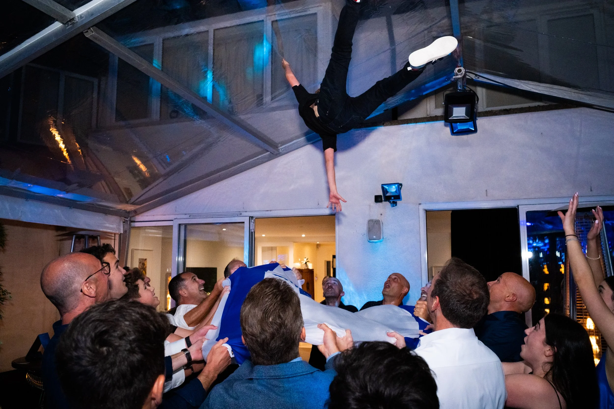 People gathered indoors at a celebration, watching a performer hanging upside down from the ceiling, reaching towards the crowd, with some holding a piece of fabric or tarp.
