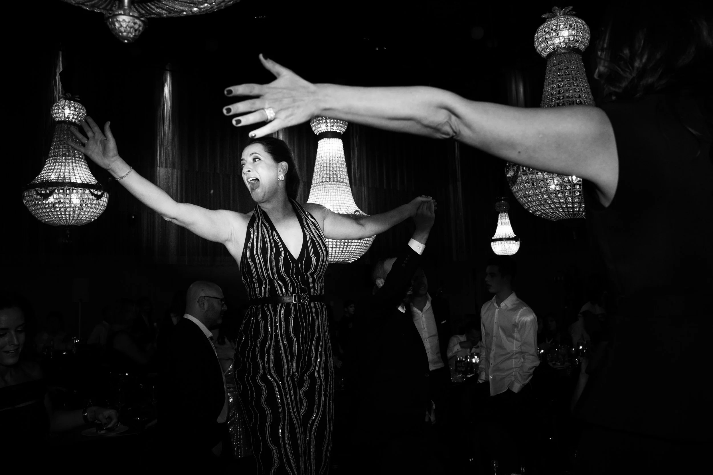 A woman in a striped dress with her arms raised and eyes closed, expressing joy, at a lively indoor event with chandeliers and other guests in the background.