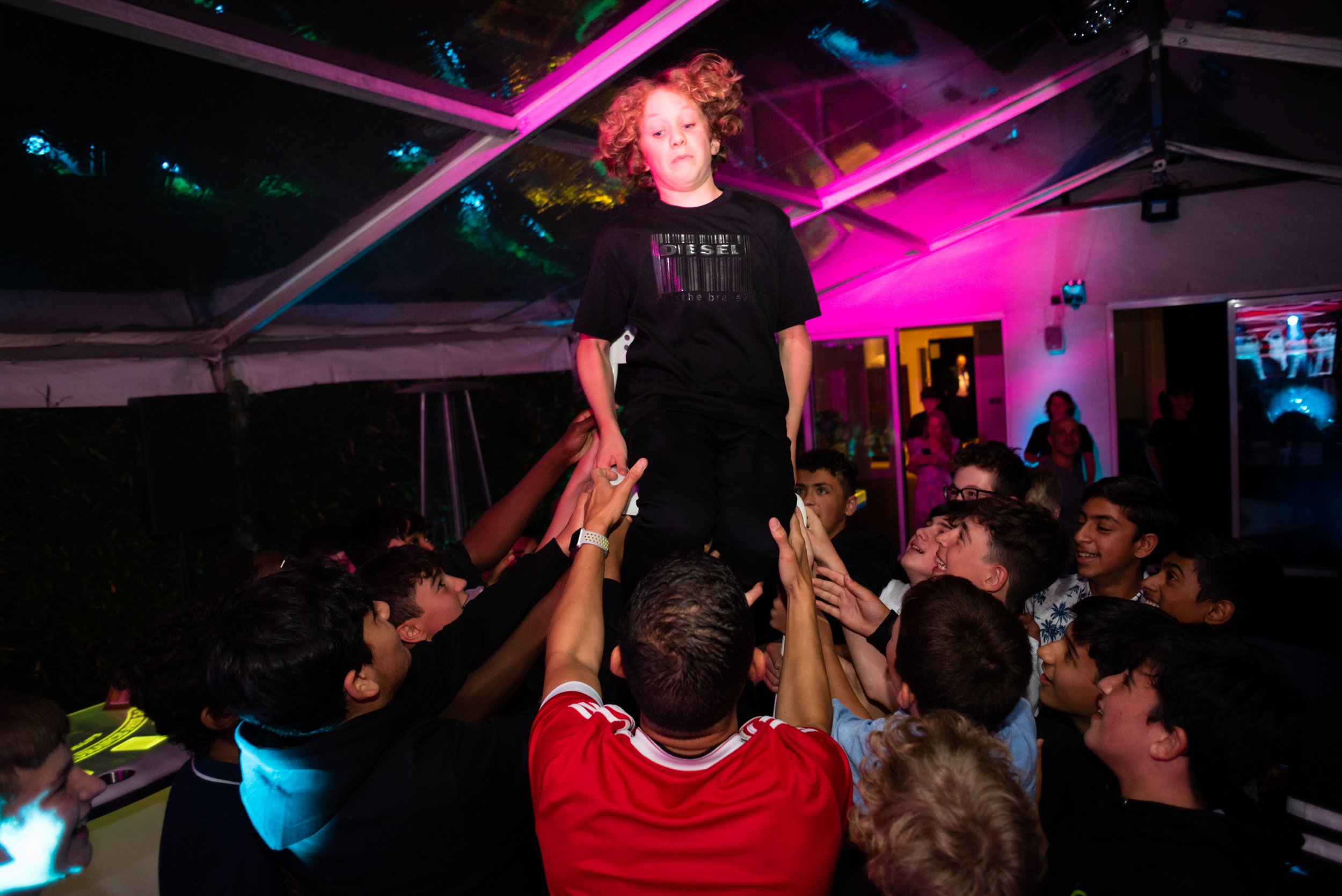 A group of kids is celebrating at a party, with some lifting a young boy on their hands in a human pyramid or support. The boy appears happy and confident, while the others are smiling and cheering. The background has colorful lighting and a tent-lik