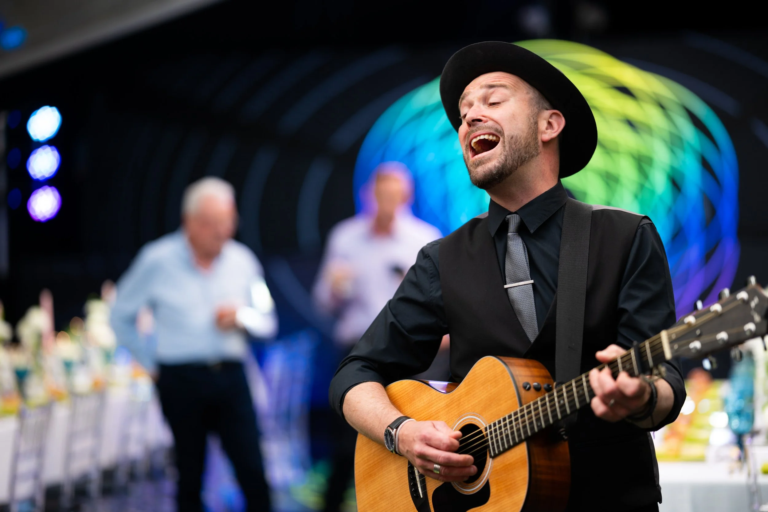 A man in a black shirt and gray tie, wearing a black hat, playing an acoustic guitar and singing, at a festive indoor event with colorful lighting and blurred people in the background.