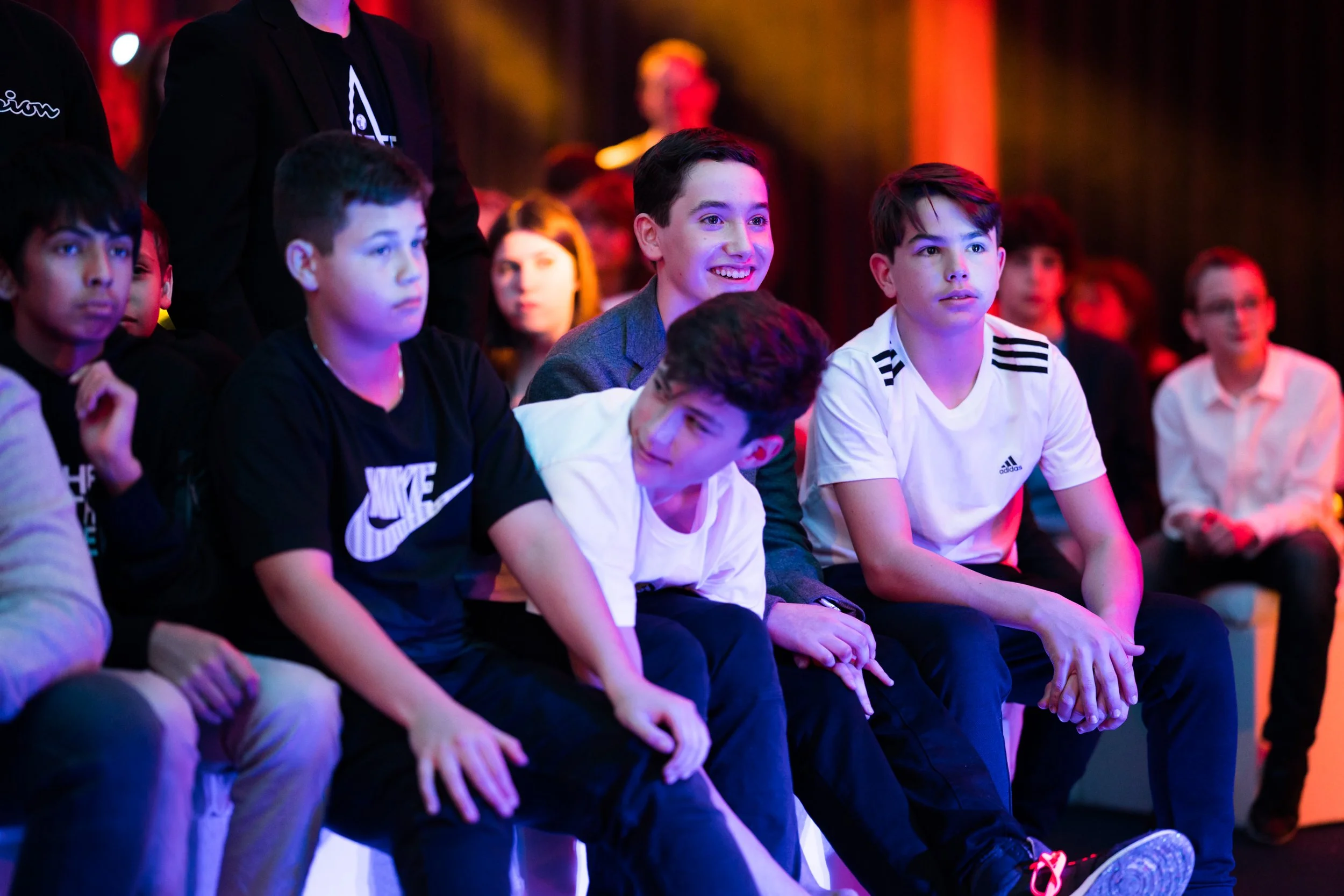 A group of boys sitting and watching an event, some are smiling, in a colorful lighting setting.