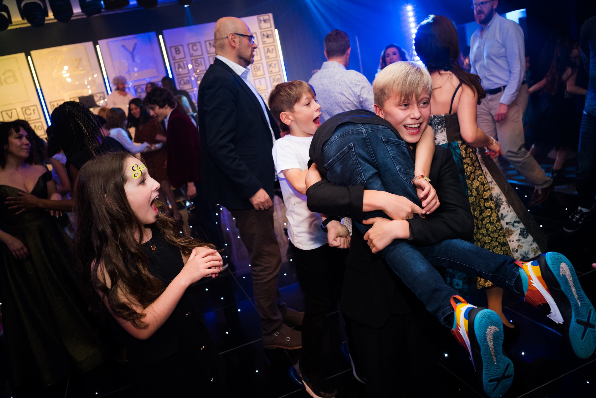 Children at a party having fun, with one boy lifting another into the air as they laugh, surrounded by other kids and adults in a decorated indoor venue.