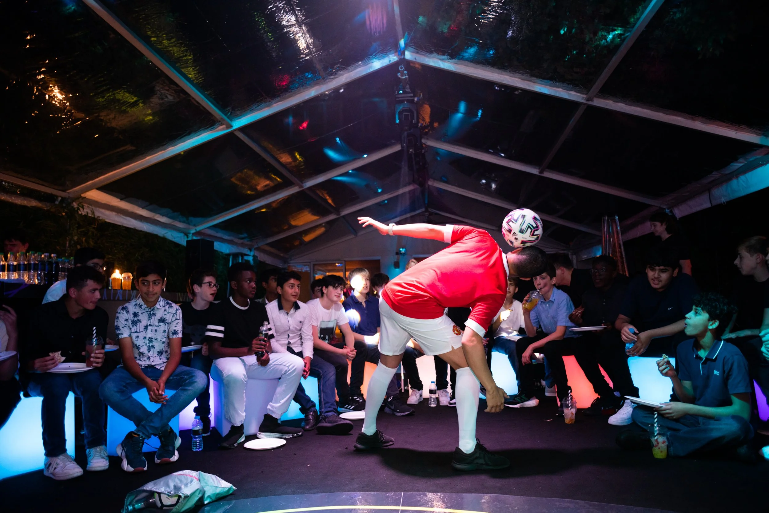 A person in a red shirt and white shorts balancing a soccer ball on their head in the center of a group of seated young people in a tent with colorful lighting.