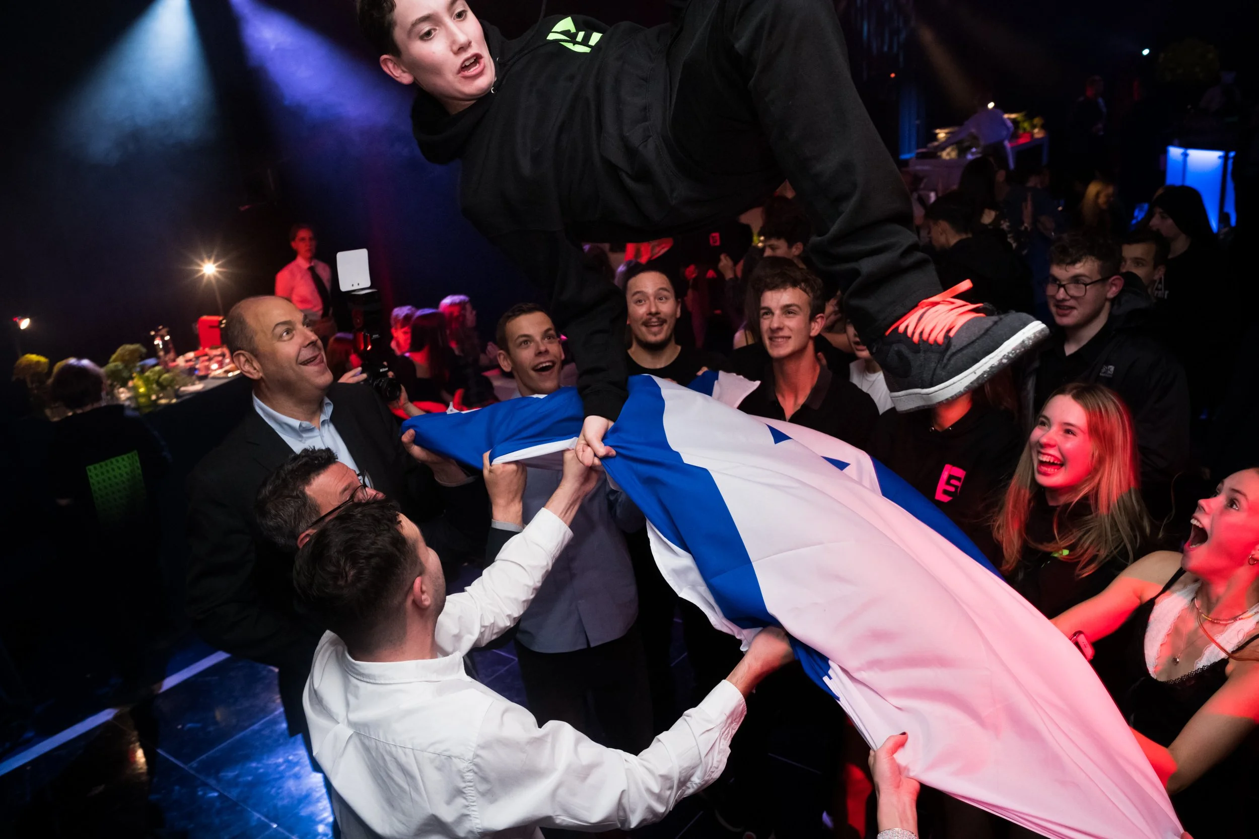 A group of people at an event celebrating, lifting a flag while a person in a black hoodie is being hoisted above them. The scene has colorful lighting and a lively atmosphere.