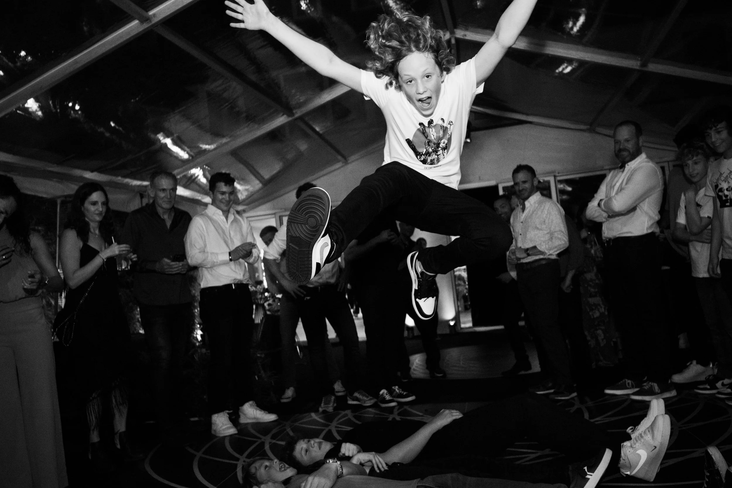 A young person is captured mid-air performing a jump at a party or event, with their arms outstretched and mouth open, over two people lying on the ground. Several people are watching and smiling in the background inside a tent or indoor venue.