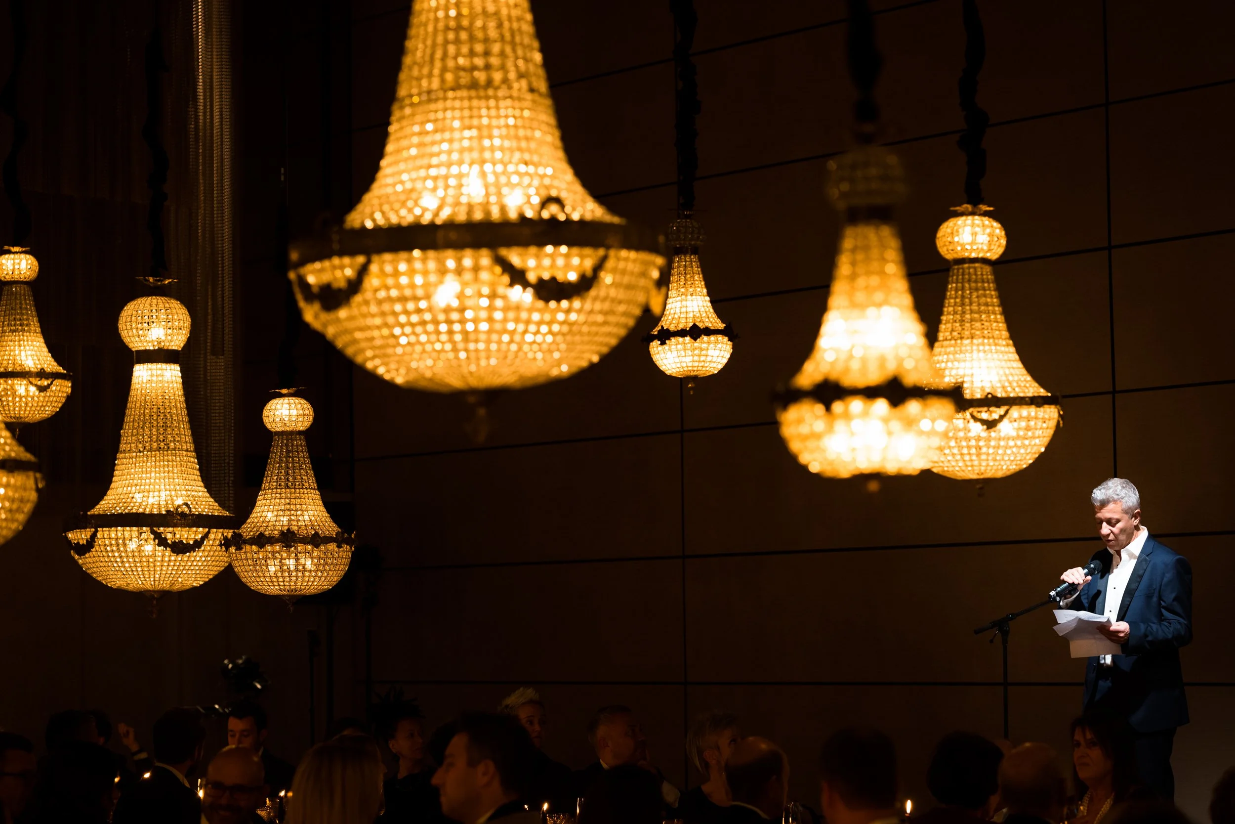 Man in tuxedo giving speech on stage at a formal event, with large ornate chandeliers hanging from the ceiling overhead, and an audience seated at tables in the background.