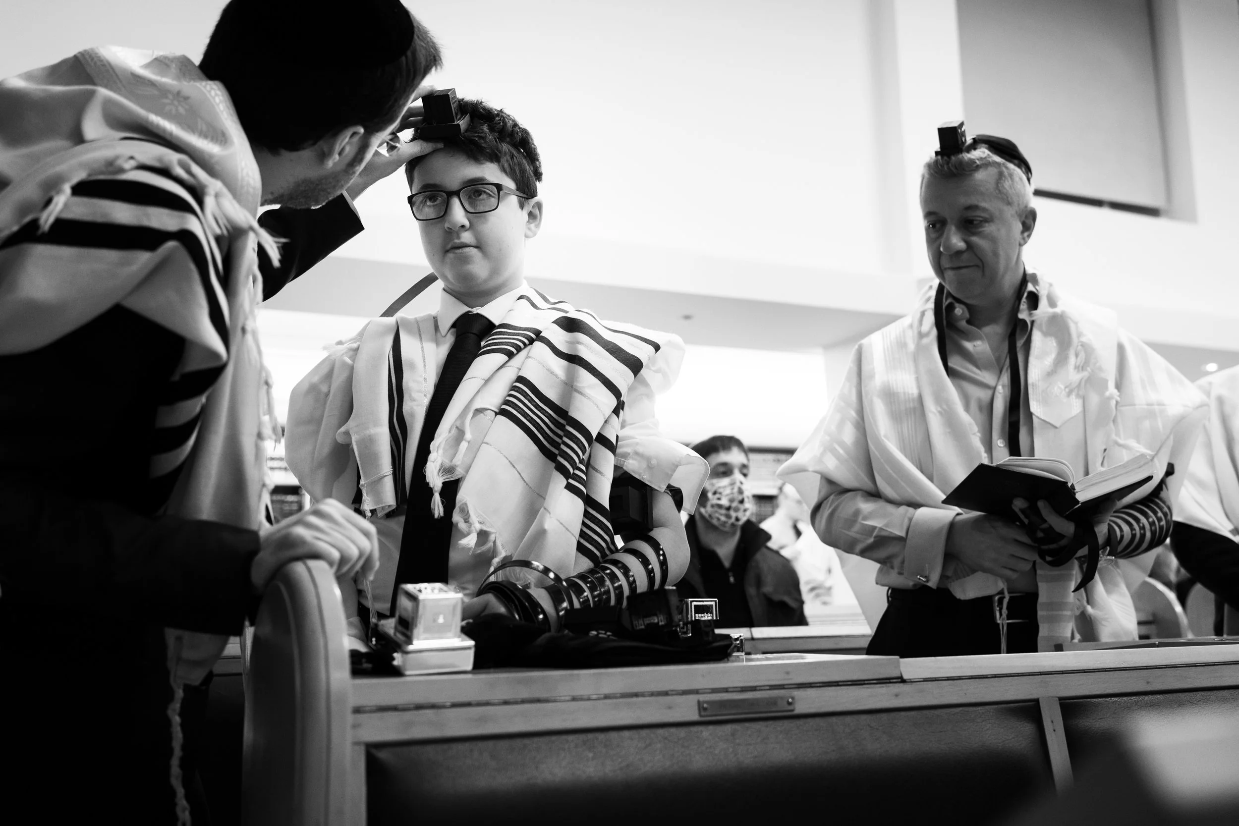 Young boy wearing glasses, a tallit, and tefillin being examined by a man with a beard and another man holding a book, all in a synagogue setting.