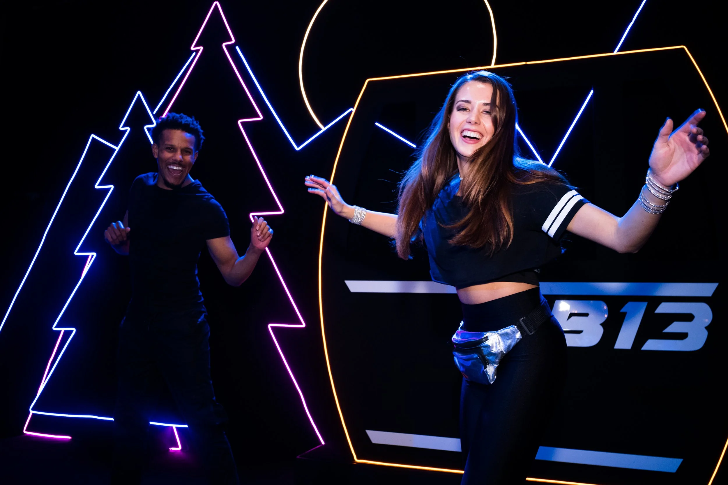 Two smiling people dancing in front of neon lightning bolt and circle shapes with a digital display in the background.