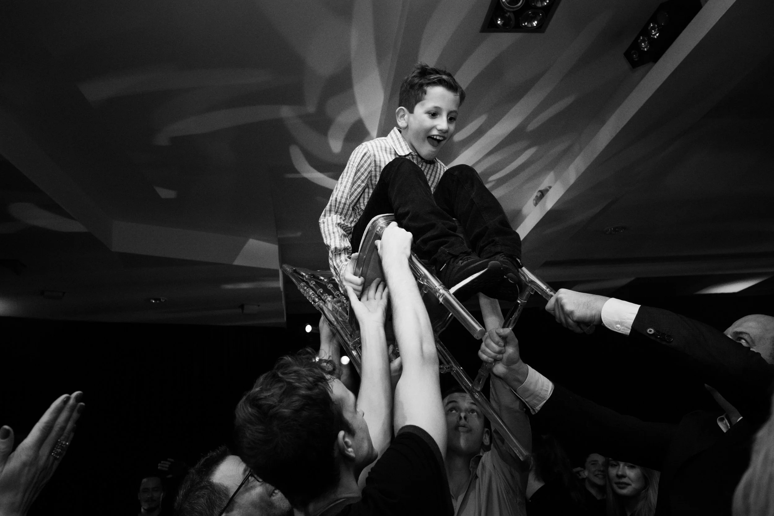 A boy being lifted on a chair at a celebration or party, with people around him holding the chair, smiling and cheering.