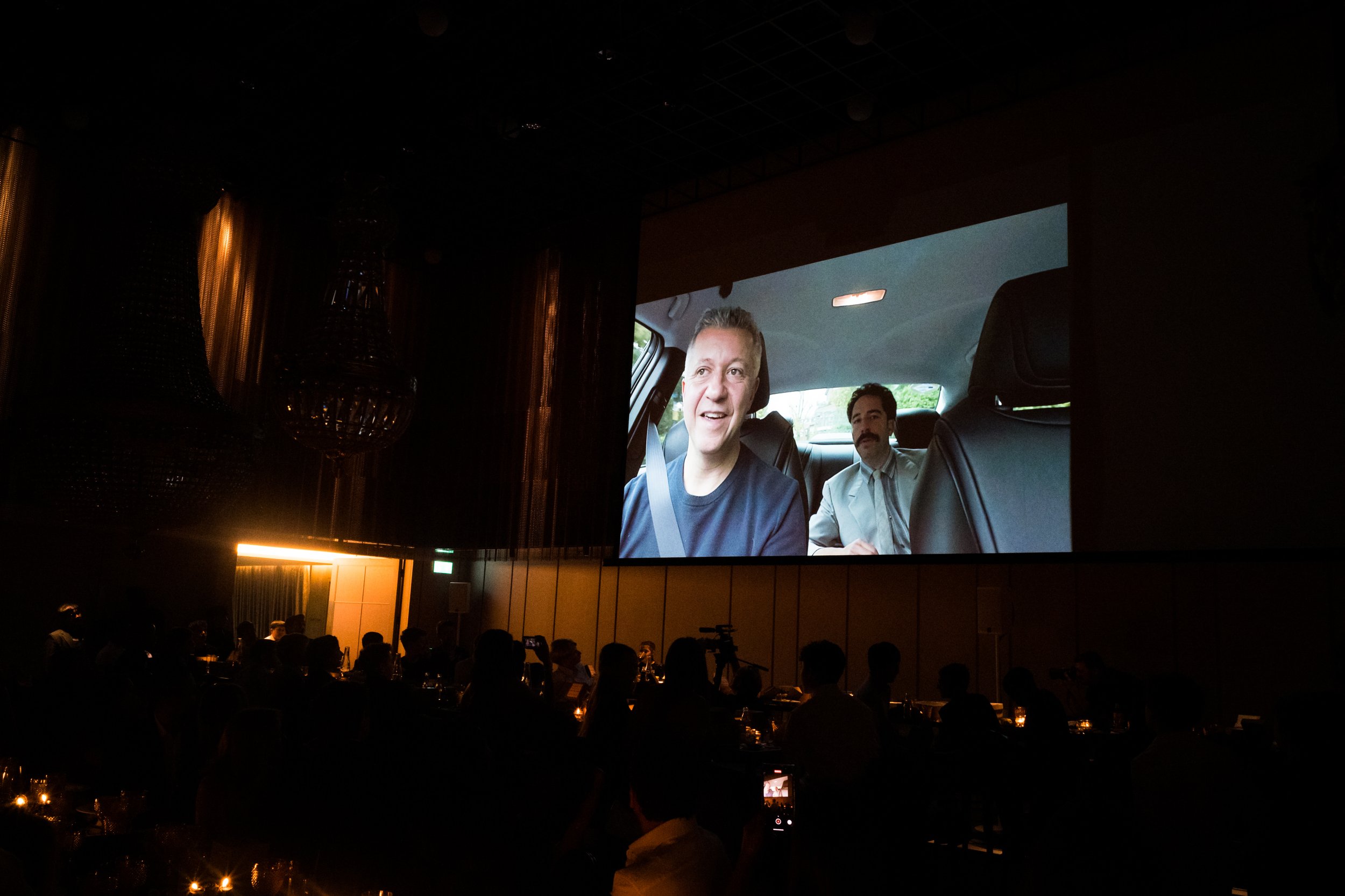 People watching a movie or presentation projected on a large screen in a dark room, with silhouetted audience members.