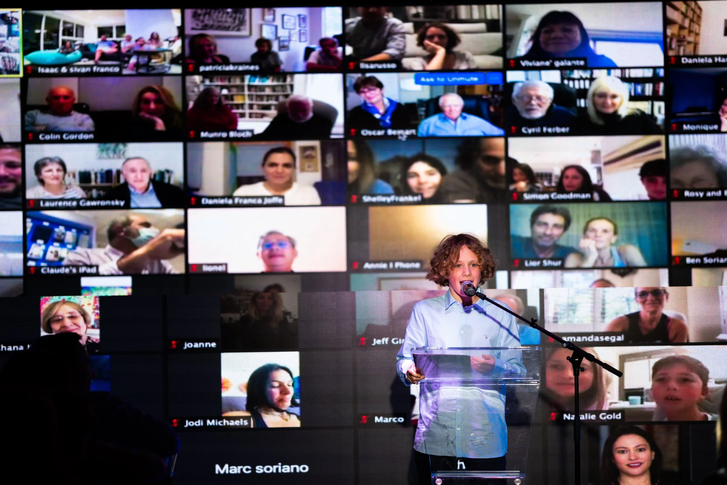 A young boy with curly hair stands at a podium with a microphone, speaking during a virtual event. Behind him, a large screen displays multiple grid views of people attending via video conference, each in separate windows.