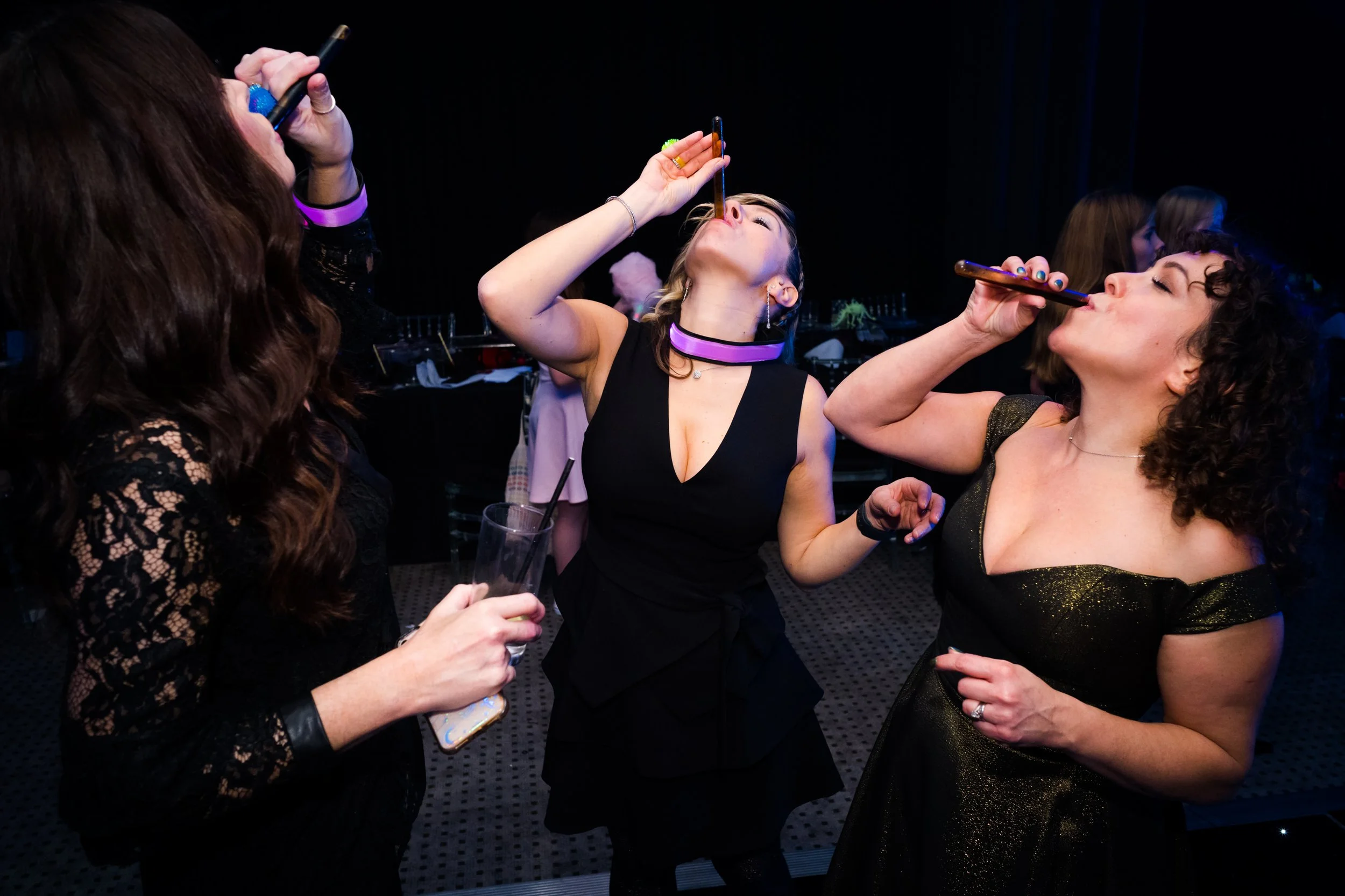 Three women in black dresses celebrating and sharing lipstick shots at a party.