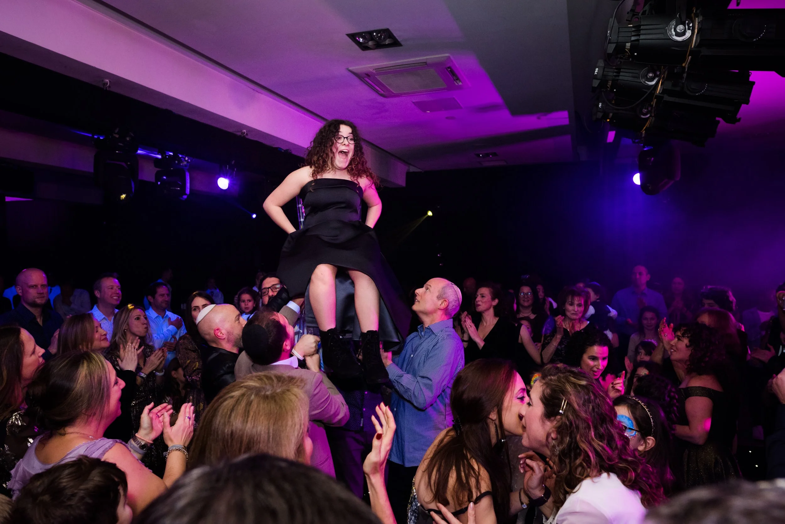 A woman in a black dress being lifted above a crowd at a party or celebration, with many people clapping and cheering, under purple and blue lighting.