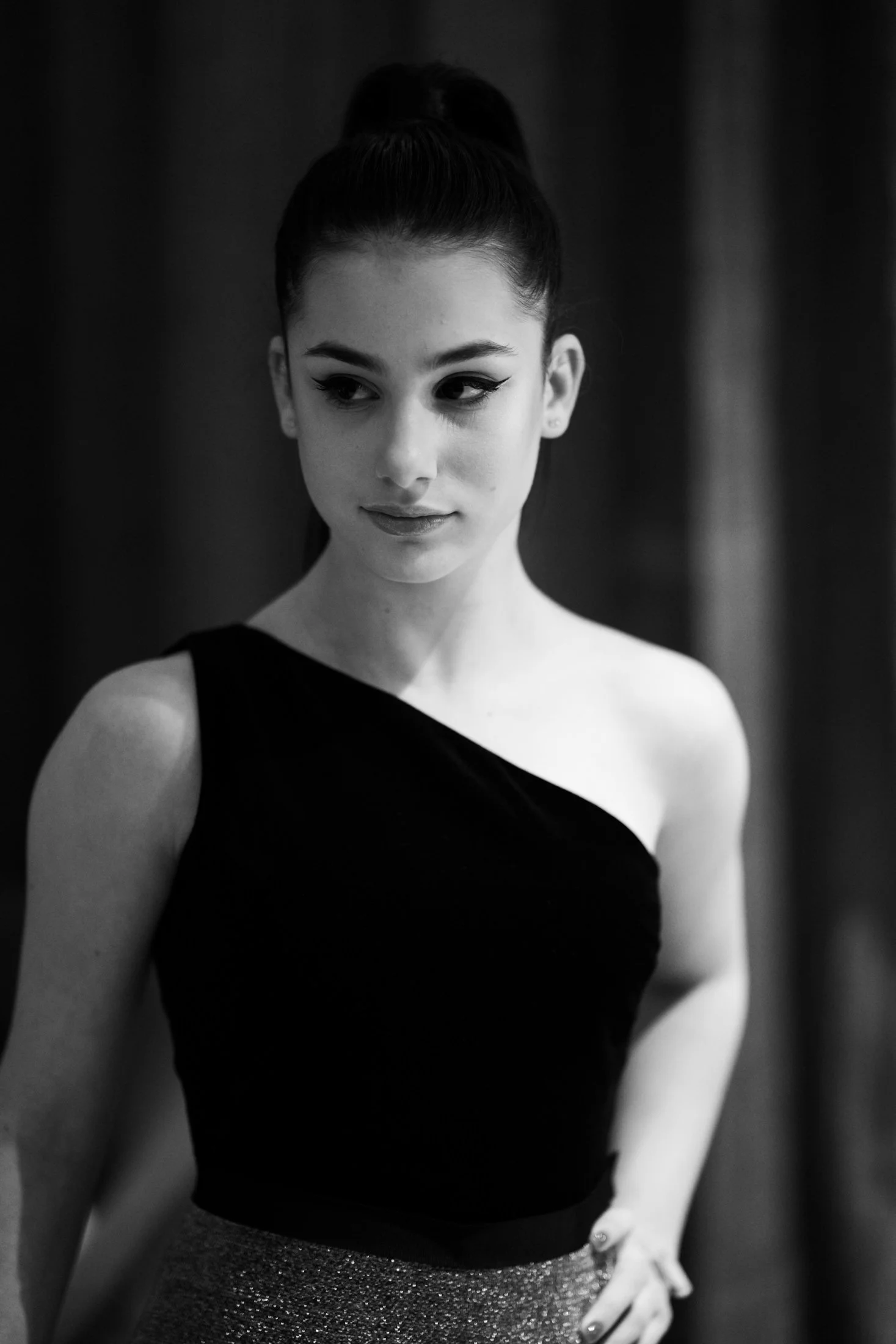 Black and white portrait of a young woman with dark hair in a bun, wearing a one-shoulder top and a sparkly skirt, posing with her hand on her hip.