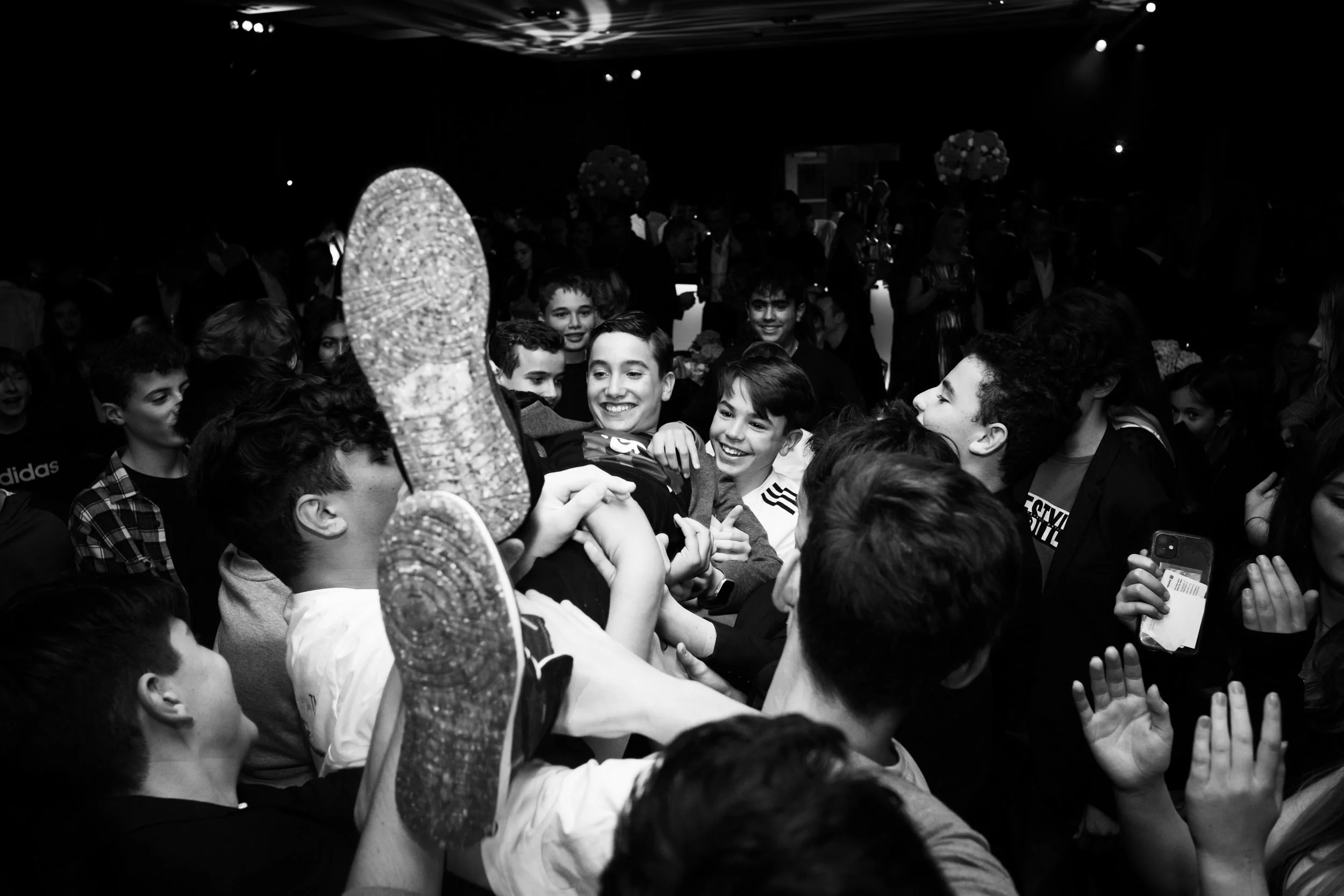 Group of young people smiling and laughing as they lift a person in the air at a crowded party or celebration.
