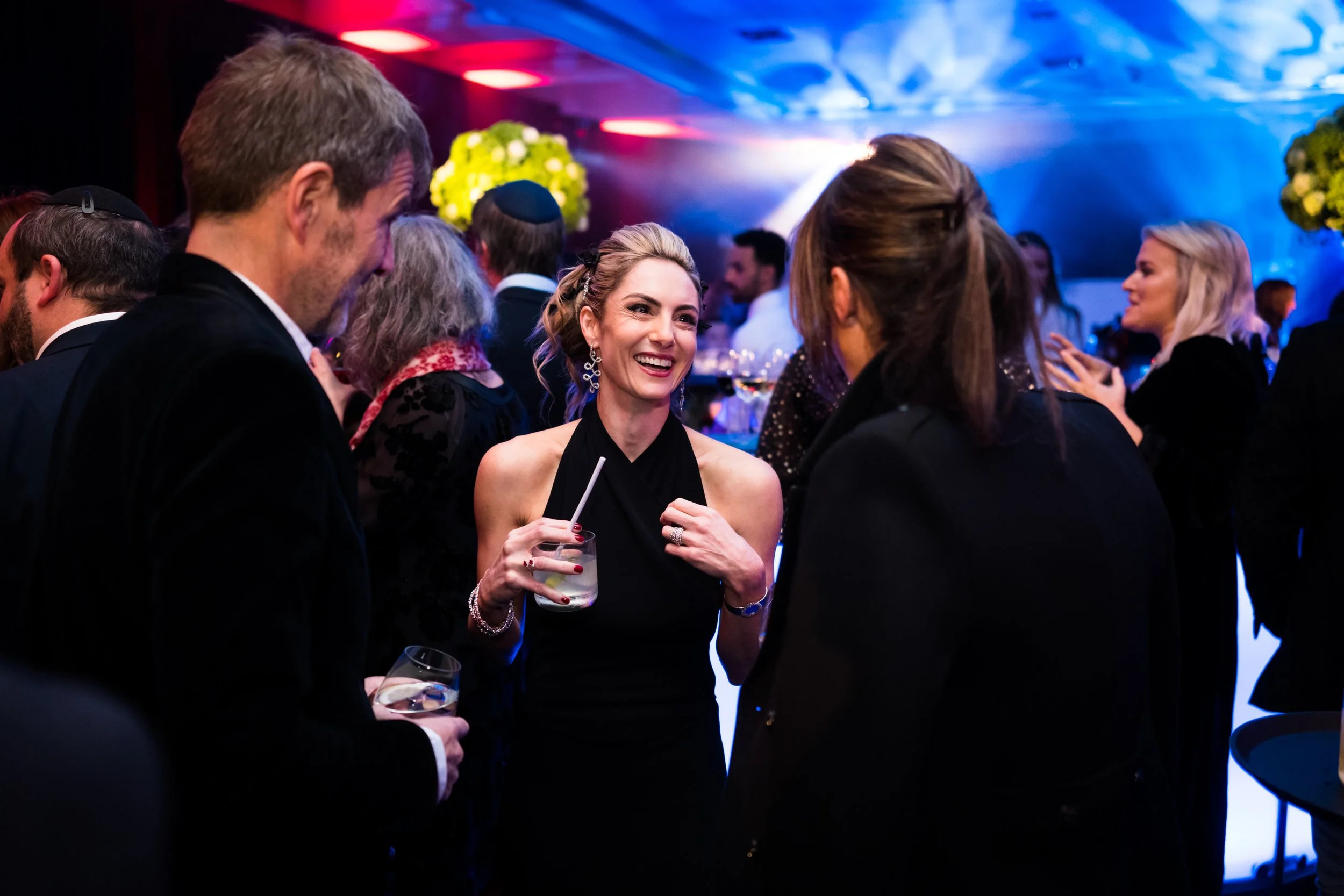 Group of people at a social event with a woman in a black dress smiling and holding a drink, engaged in conversation in a dimly lit setting with colorful lighting and floral decorations.