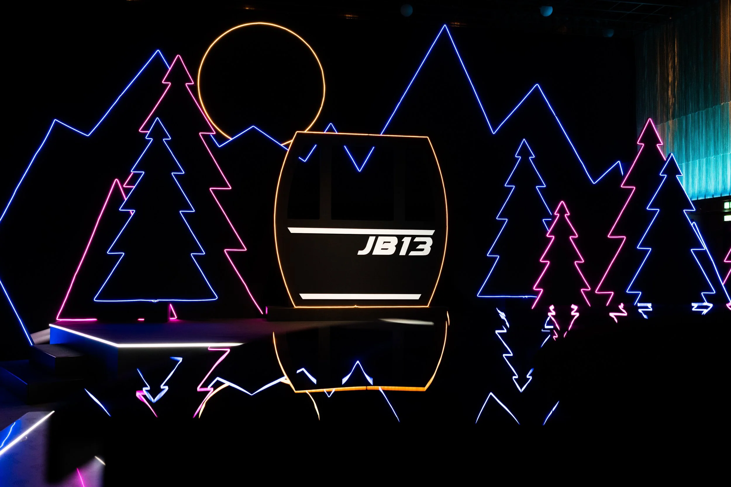 Neon mountain landscape with pine trees, a bright yellow sun, and a ski lift with 'JB13' logo, reflected in a body of water.