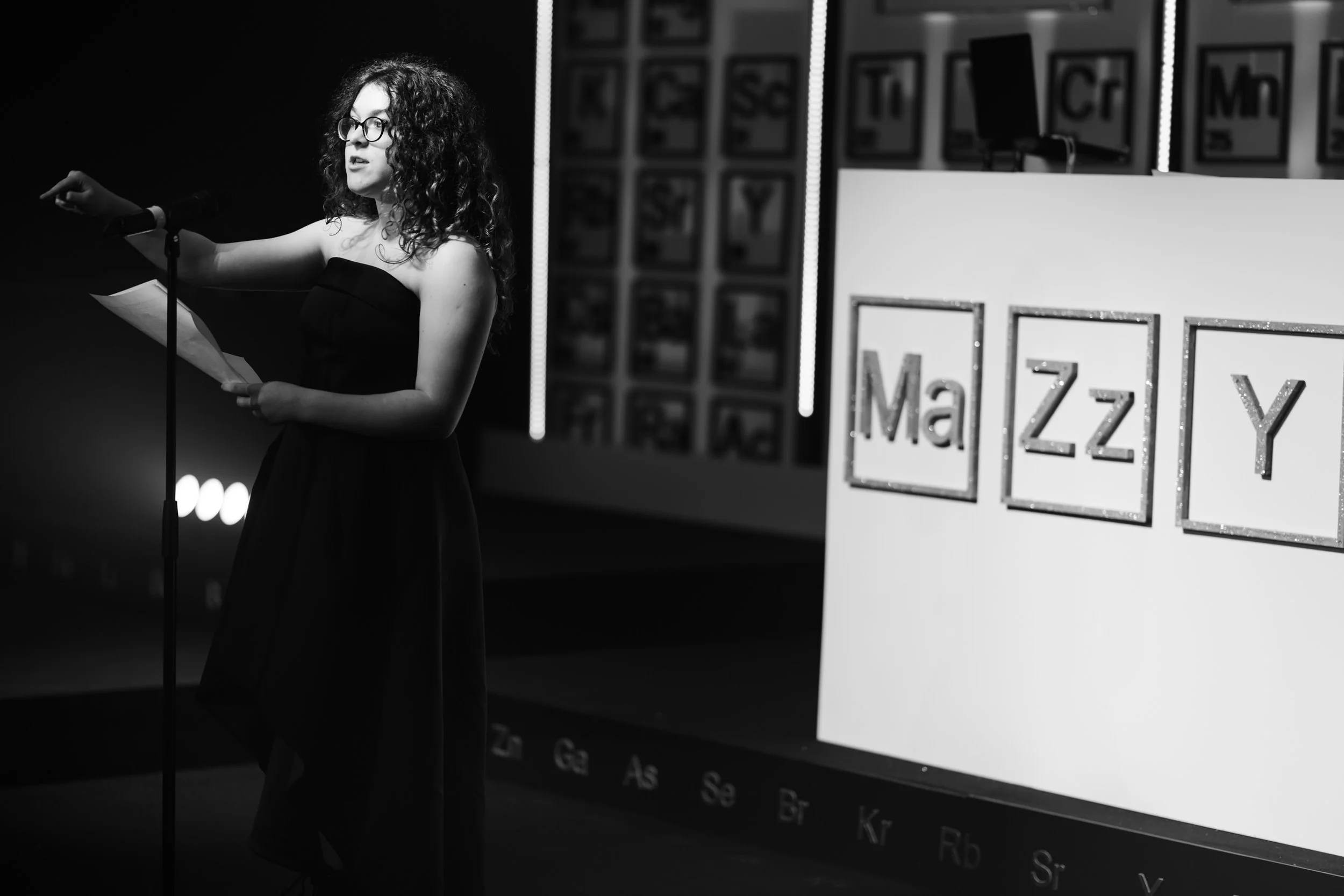 A woman with curly hair and glasses standing at a microphone, reading. She is wearing a strapless dress, and there is a large display with partial chemistry element symbols behind her.