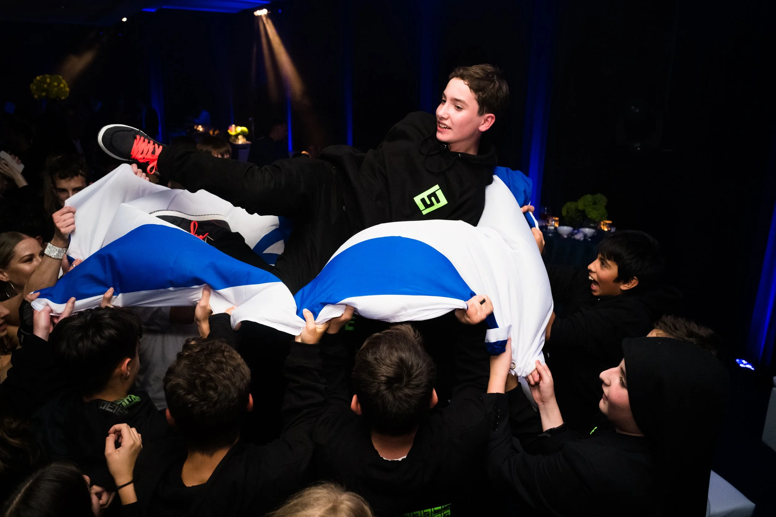 A young man being lifted on a group of people's arms during a celebration event, surrounded by excited onlookers in a dimly lit room.