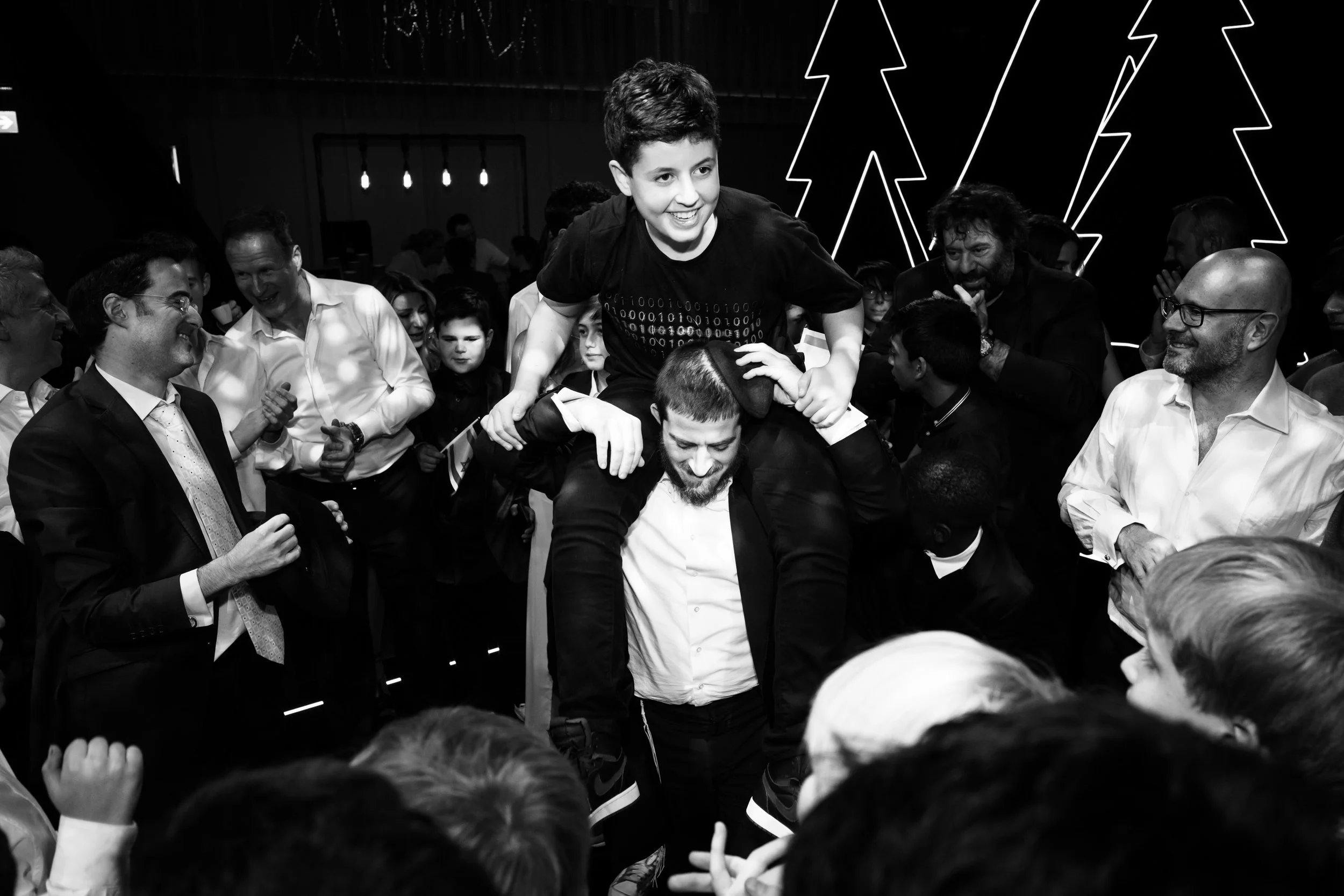 A group of people, including children and adults, are gathered at a celebration or event. A young boy is sitting on a man's shoulders, smiling, as he is part of a crowd that is cheering and enjoying the moment. The photo is in black and white.