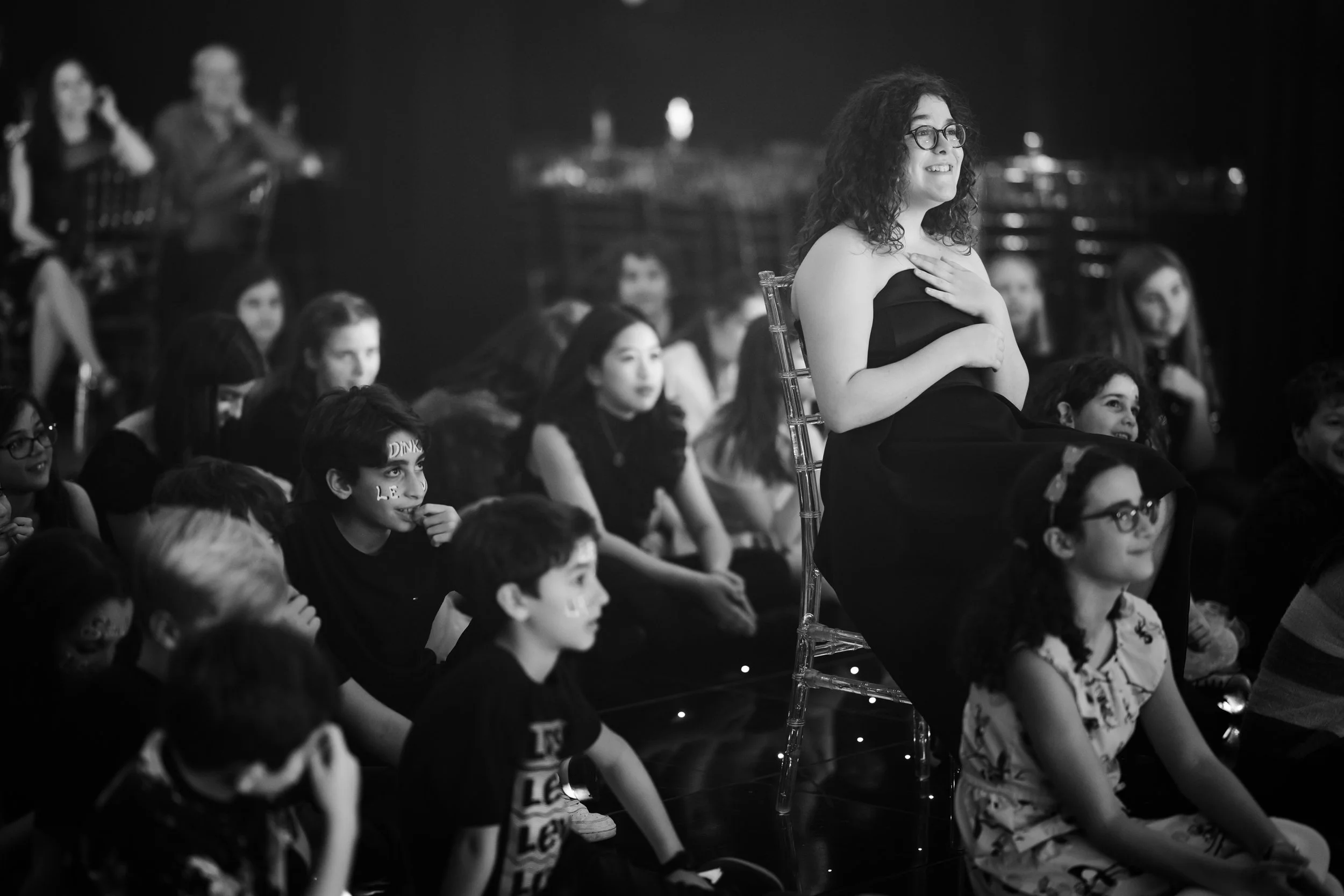 A woman in a strapless dress and glasses is sitting on a transparent chair, smiling and holding her chest with one hand, while a group of children are sitting on the floor around her, some with face paint, watching attentively. The scene appears to b