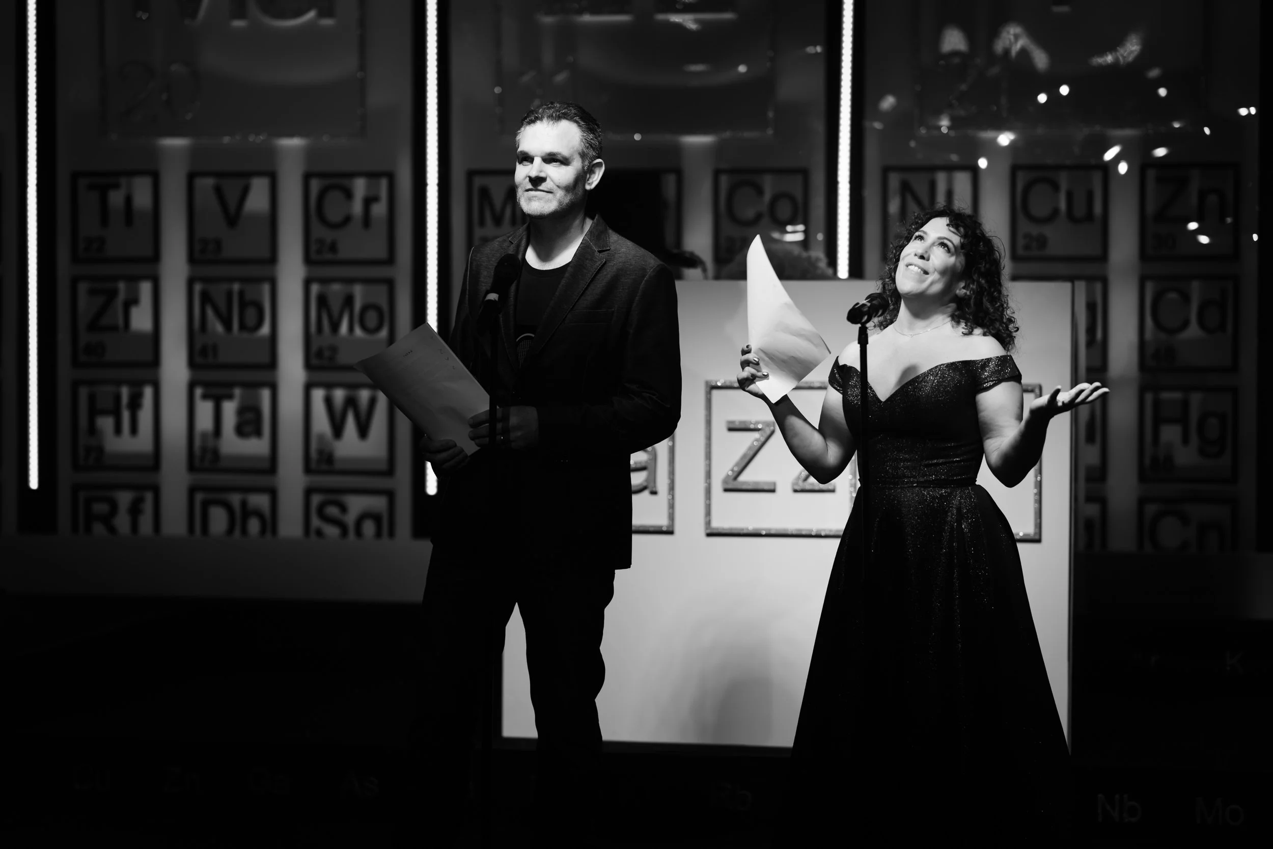 A man and woman on stage, with the man holding papers and the woman gesturing with a paper in hand. They are in front of a background with large periodic table elements.