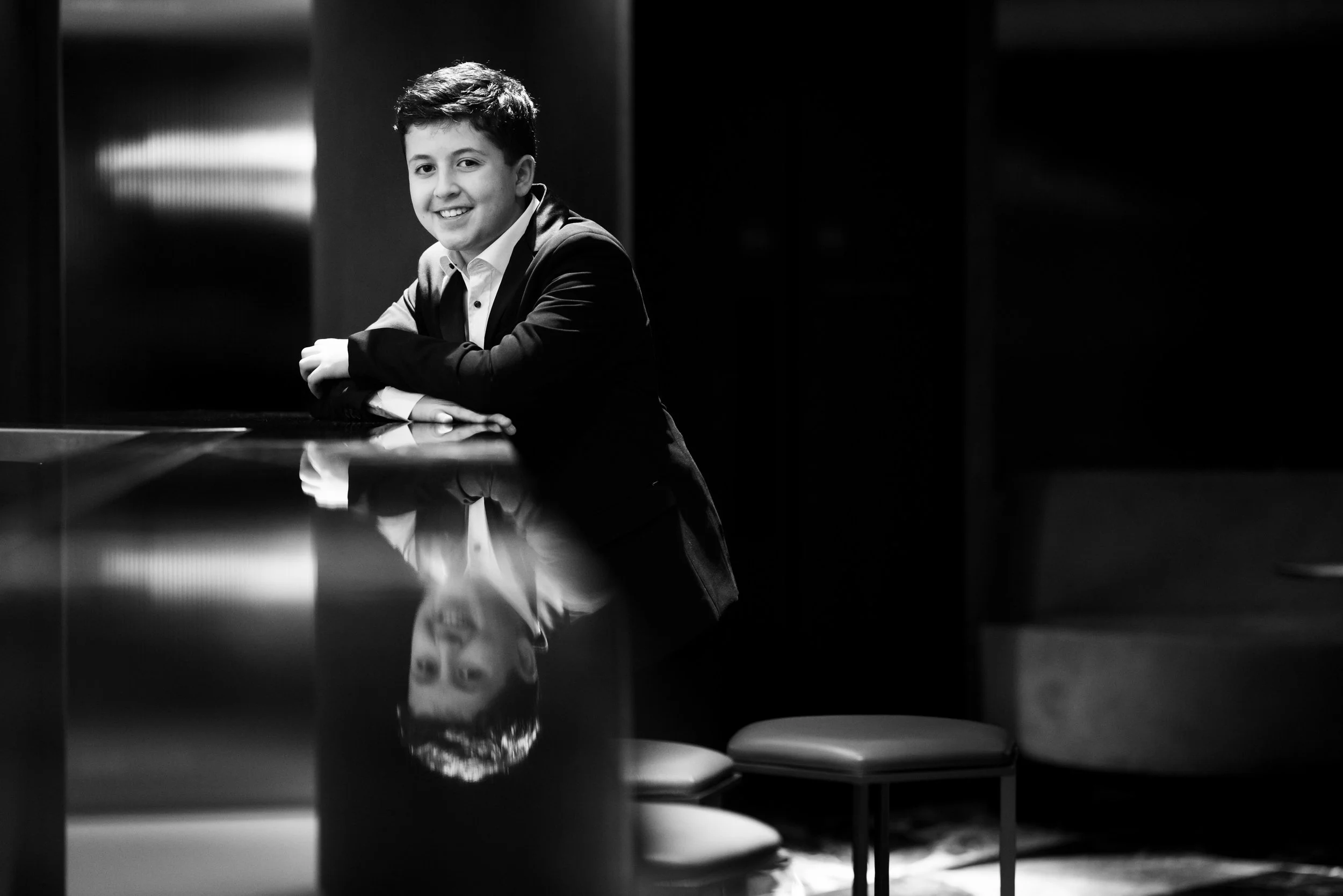 A young man in a suit sitting at a reflective table, smiling, with his reflection visible in the surface. The image is in black and white.